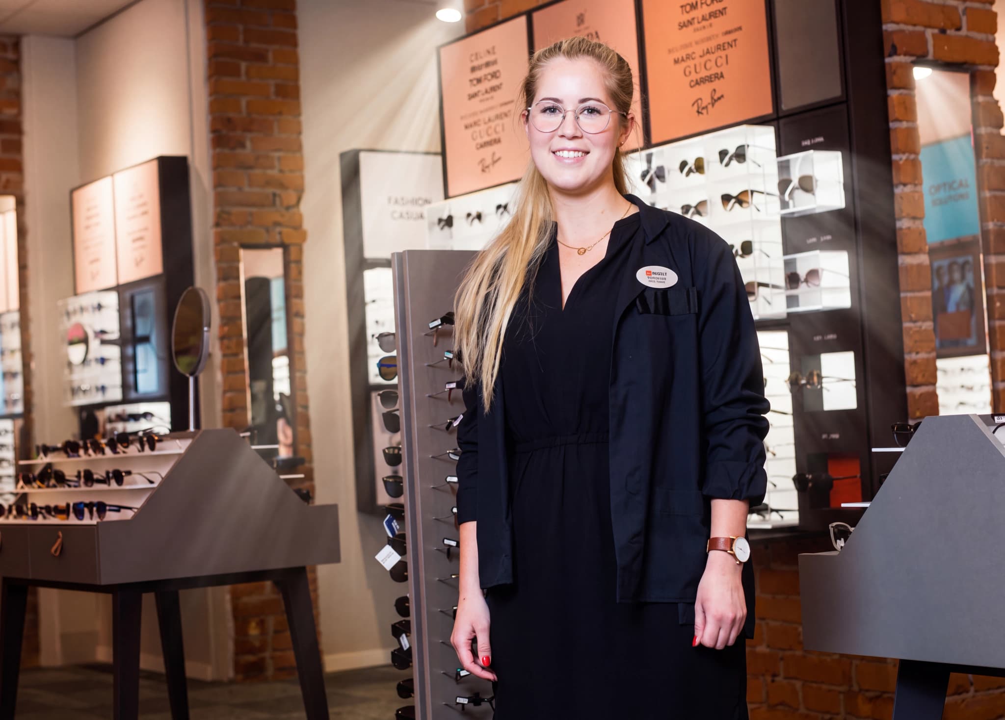 En leende ung kvinna som arbetar som optiker står i en butik med glasögon och solglasögon uppradade.