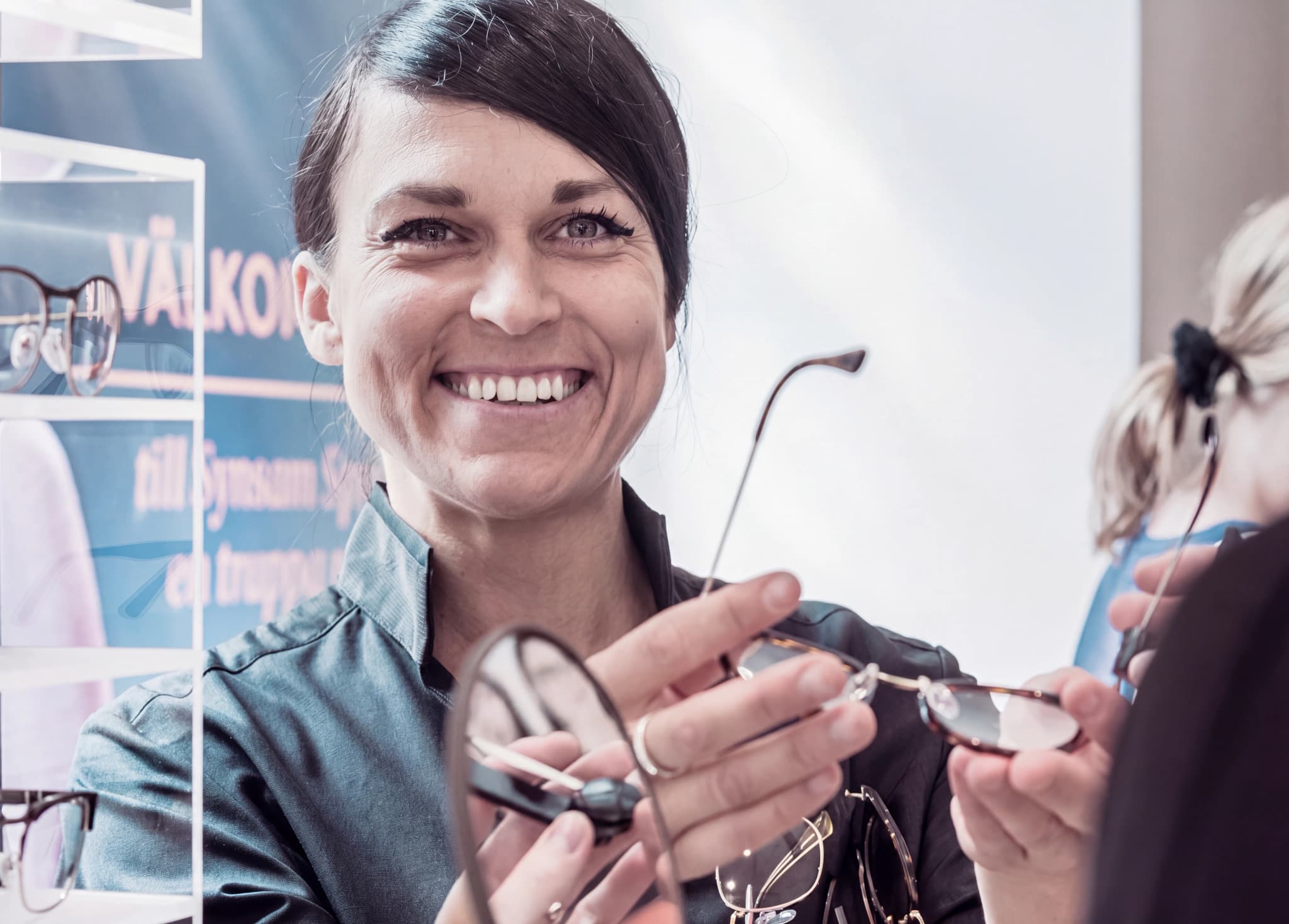 En optiker ler och visar upp ett par glasögon för en kund i en butik, med glasögon utställda i bakgrunden.