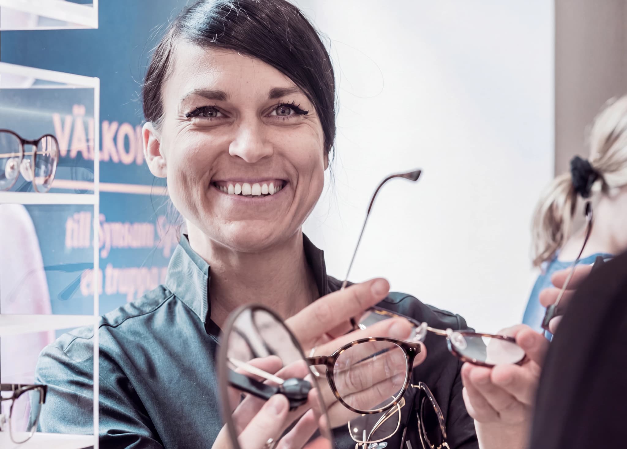 Optiker ler och visar upp ett par glasögon för en patient i bakgrunden i en butik eller mottagning.