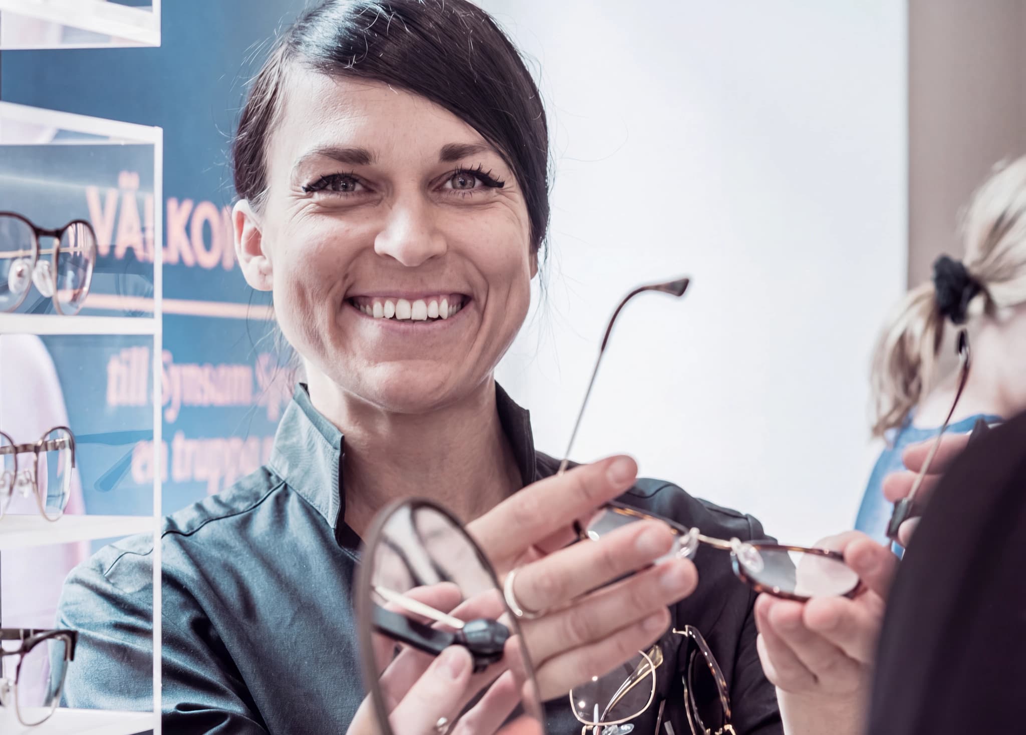 En leende optiker visar glasögon för en kund i en butik med synundersökningsutrustning.