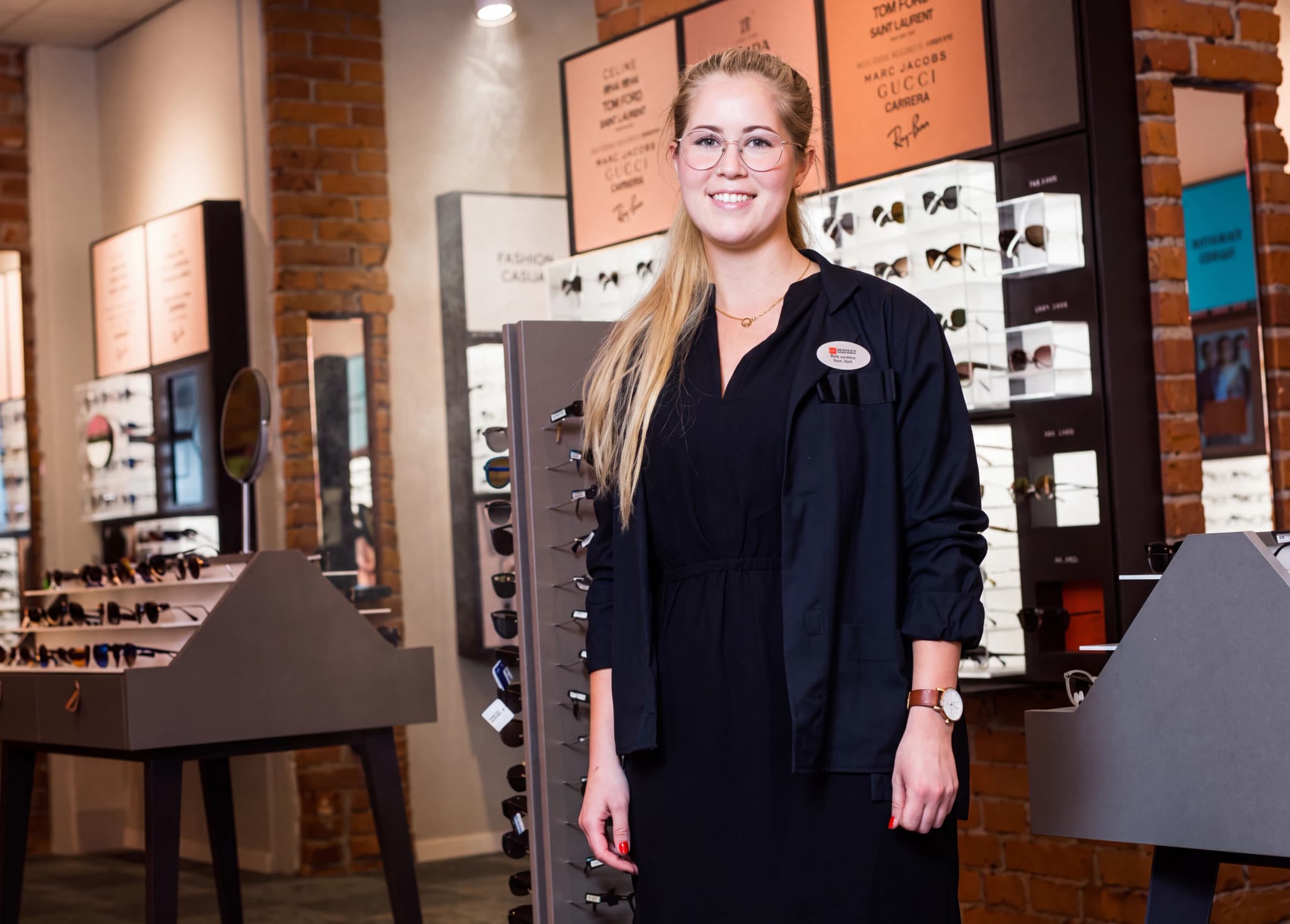 Optiker i arbetskläder ler och tittar framåt i en glasögonbutik, med glasögonställningar i bakgrunden.