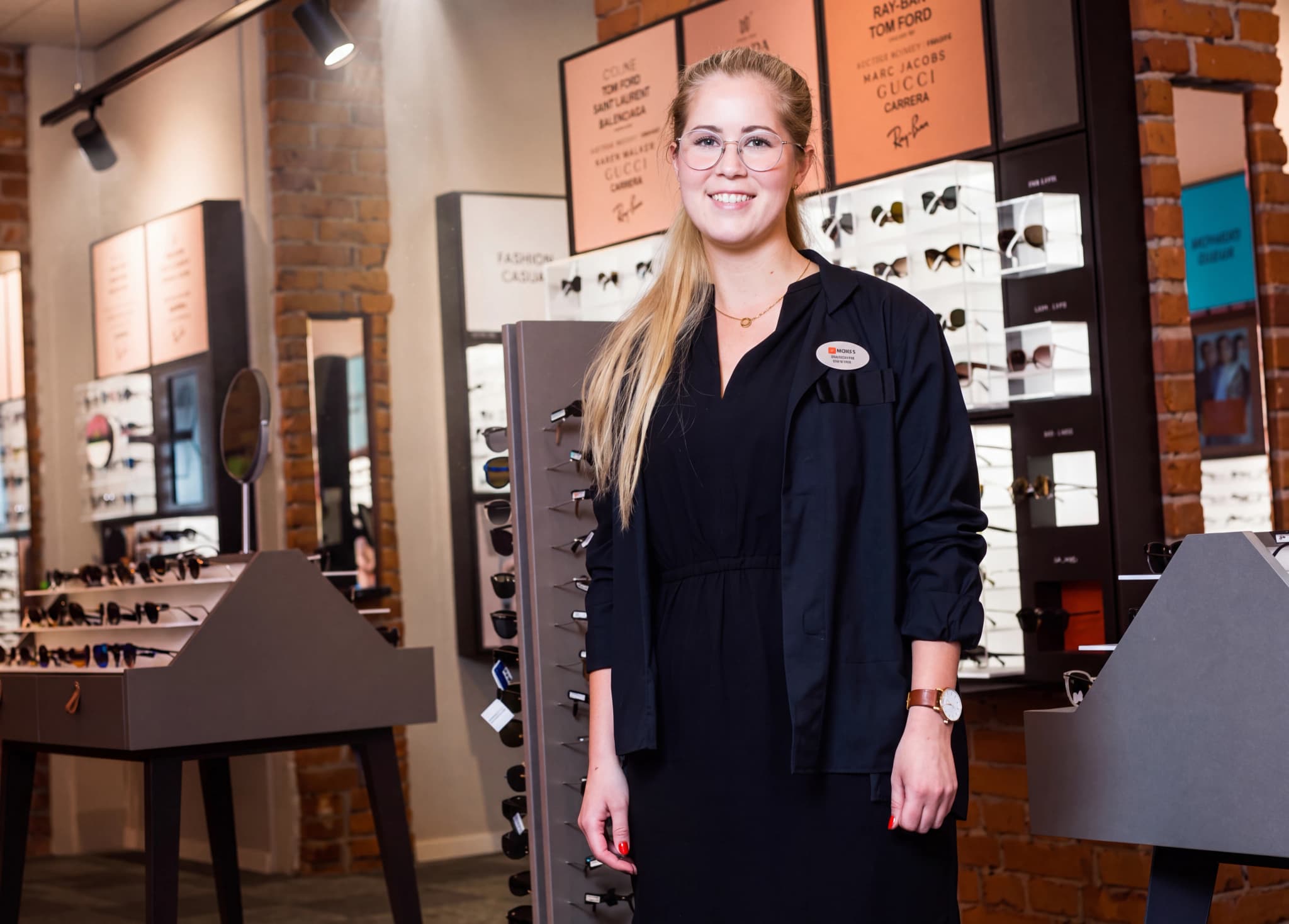 En ung kvinna med glasögon och blont hår ler mot kameran, klädd i en svart jacka och klänning. Hon står i en butik som säljer glasögon och solglasögon, med displayer av glasögon i bakgrunden.
