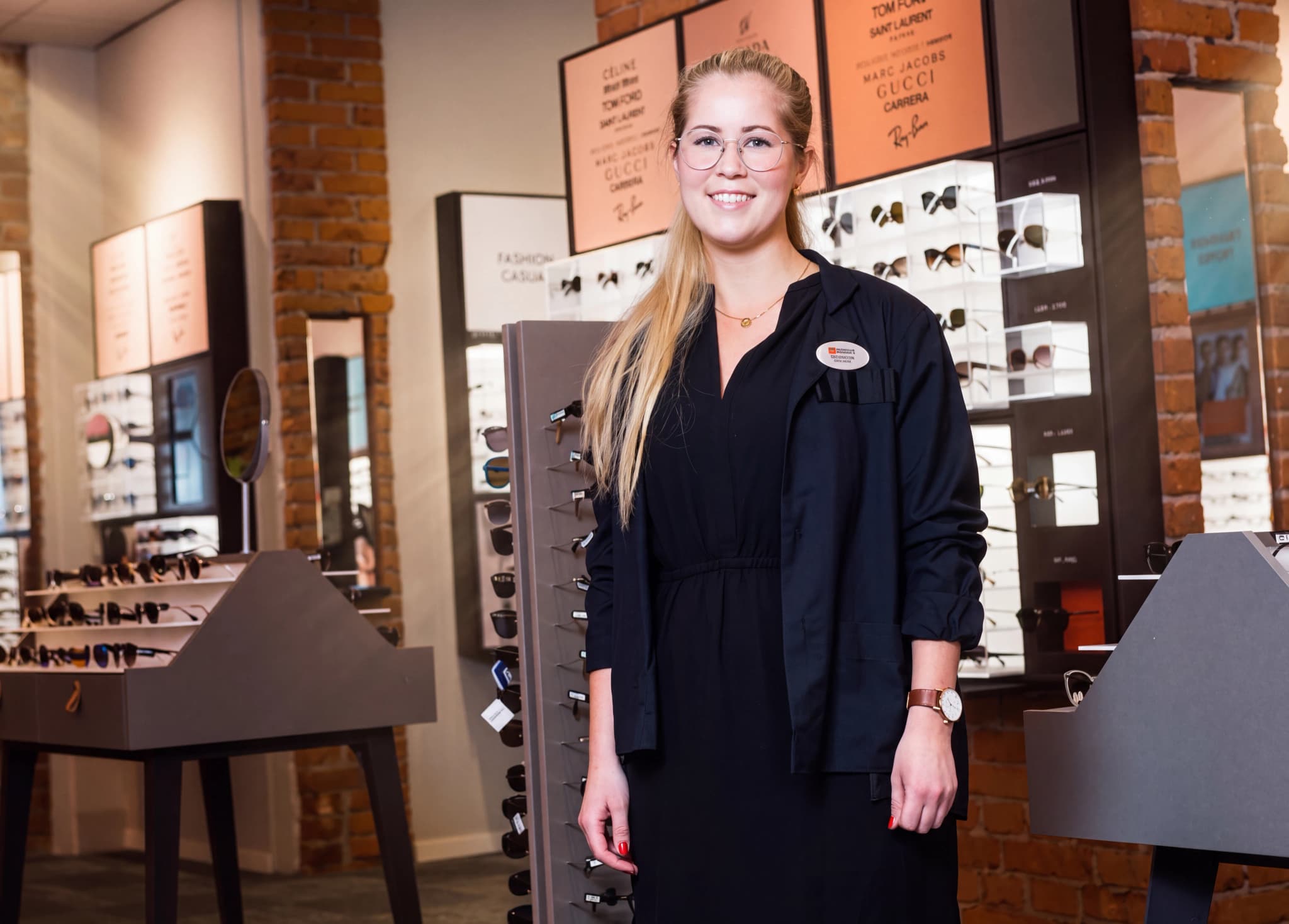 En leende ung kvinna i arbetskläder och glasögon står i en butik som säljer glasögon och solglasögon.