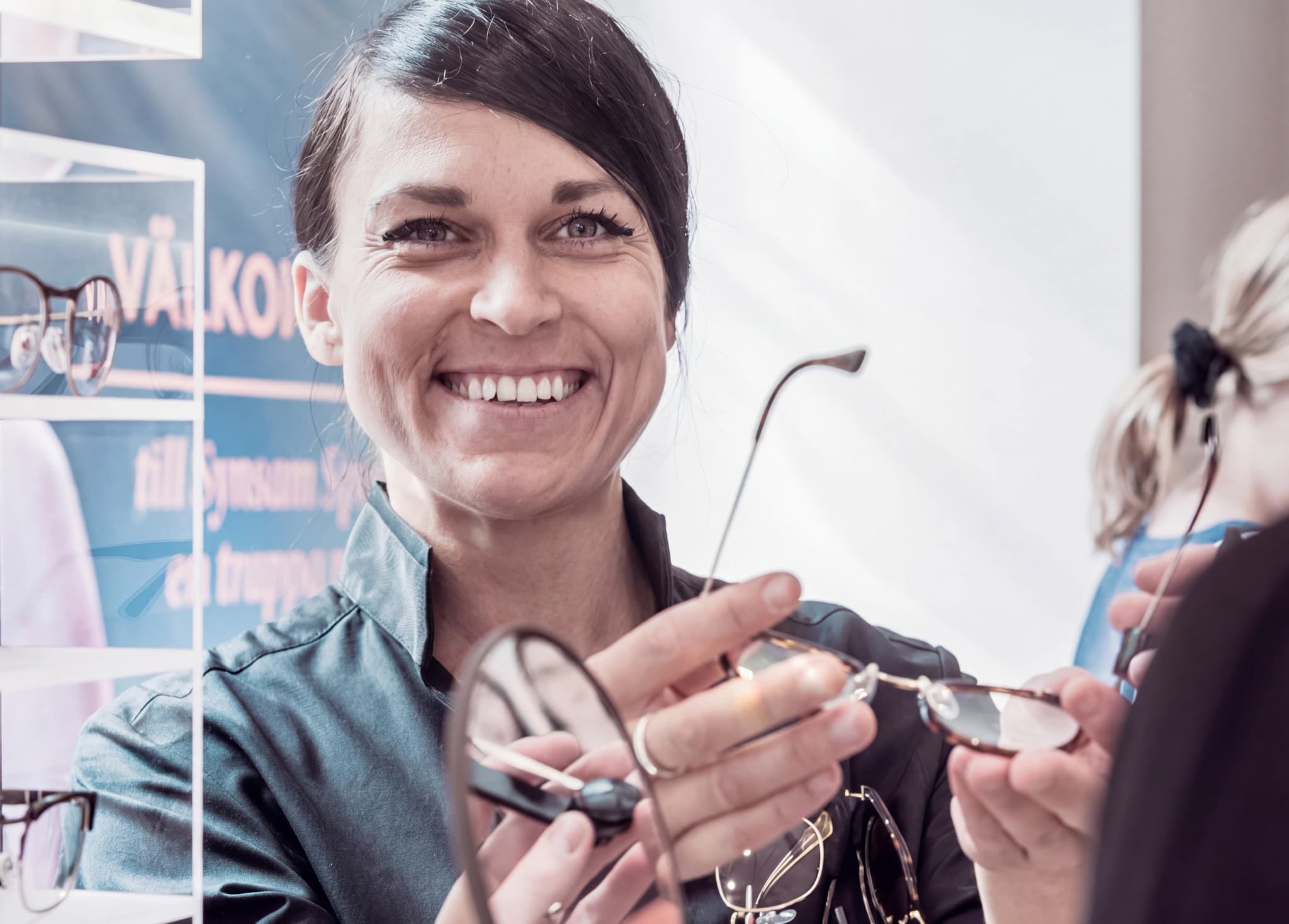 En optiker ler och visar upp ett par glasögon för en kund i butiken.