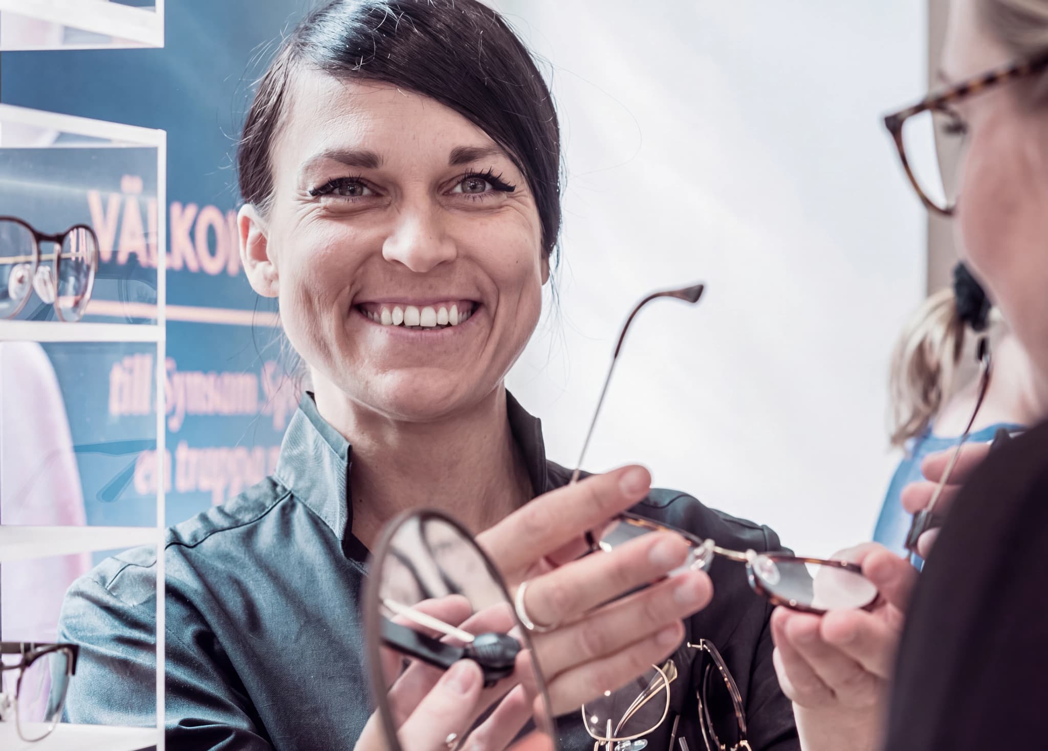 En optiker ler och visar ett par glasögon för en kund i en butik.