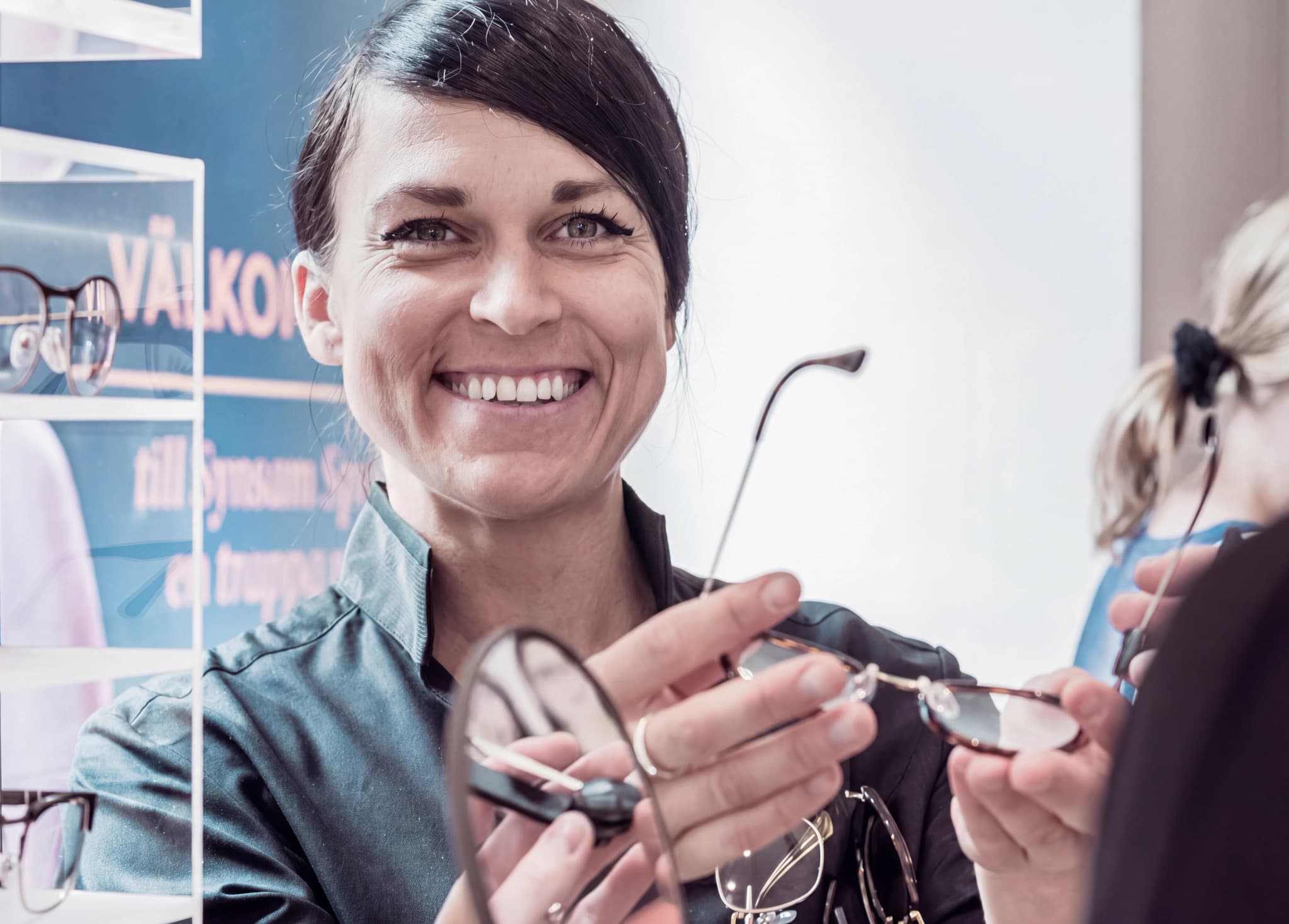 En optiker ler och håller upp ett par glasögon för en kund i en synundersökningsmottagning.
