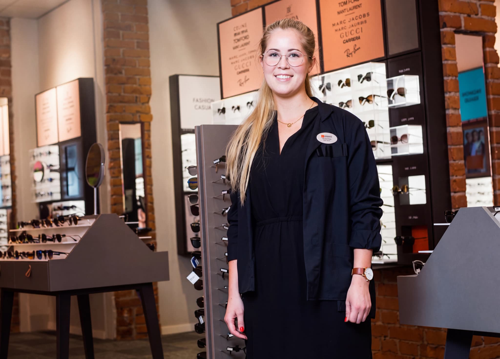 En ung kvinna med glasögon, klädd i mörka kläder och med en namnskylt, står leende i en optikerbutik omgiven av glasögonställningar.
