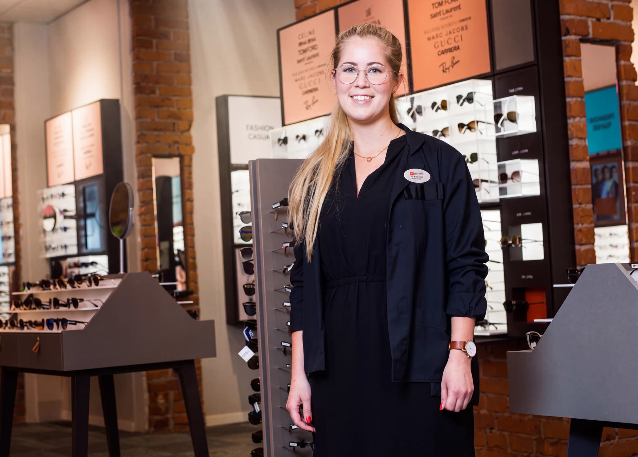 Optiker står i butik med glasögonbakgrunder och displayer.