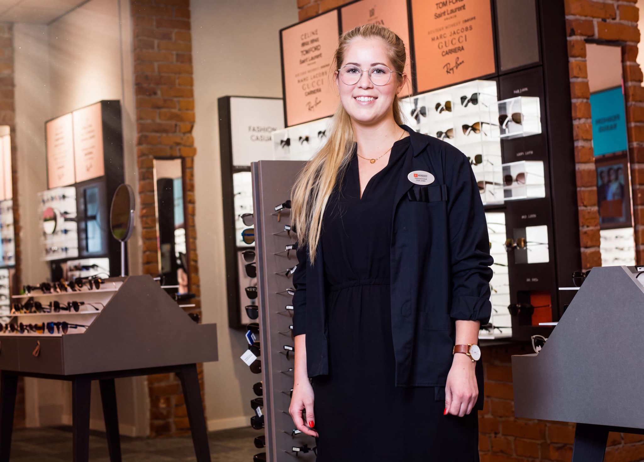 En ung kvinna som arbetar som optiker står leende i en butik med glasögonutställningar i bakgrunden.
