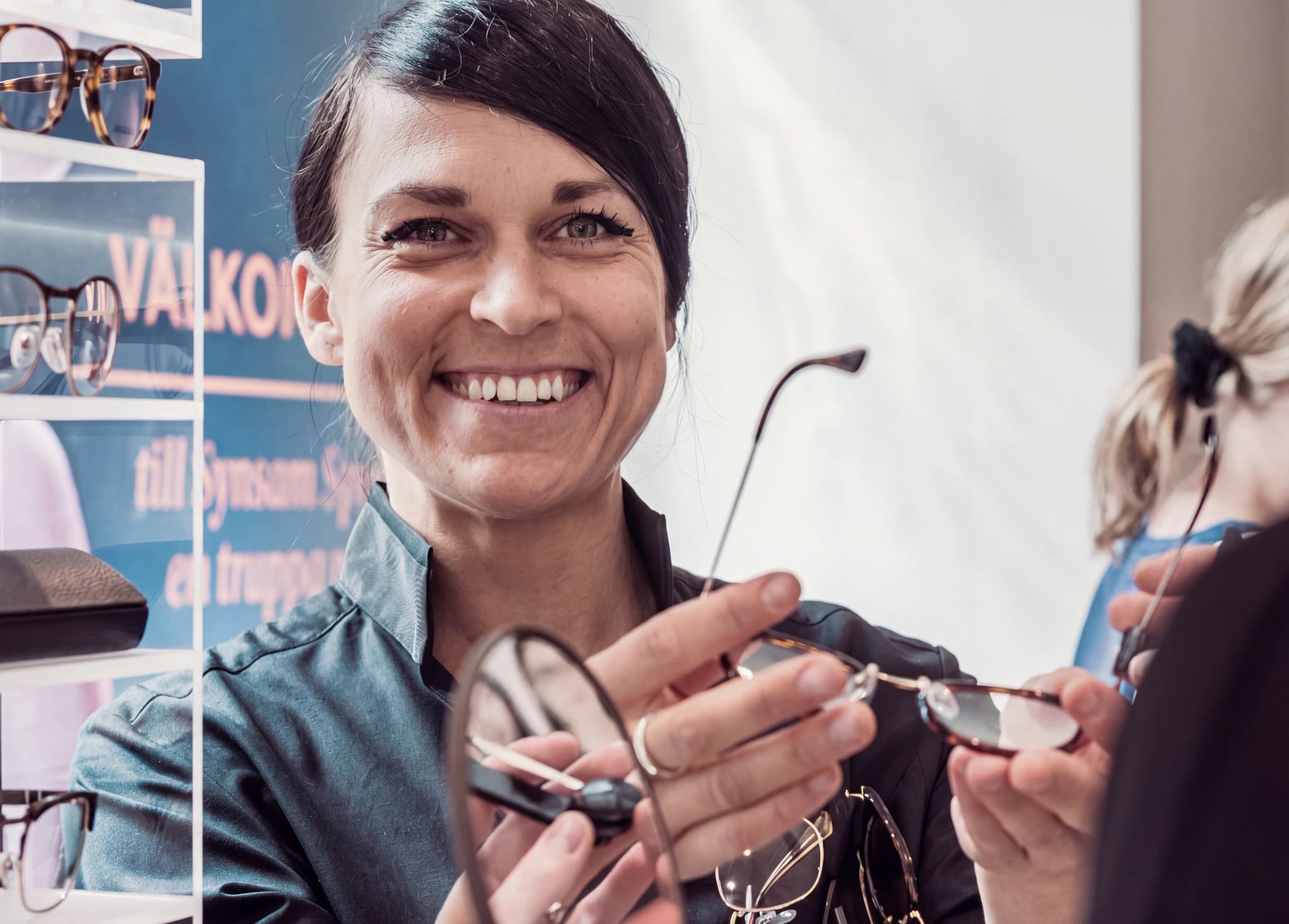 En optiker ler och visar ett par glasögon till en kund i en butik.