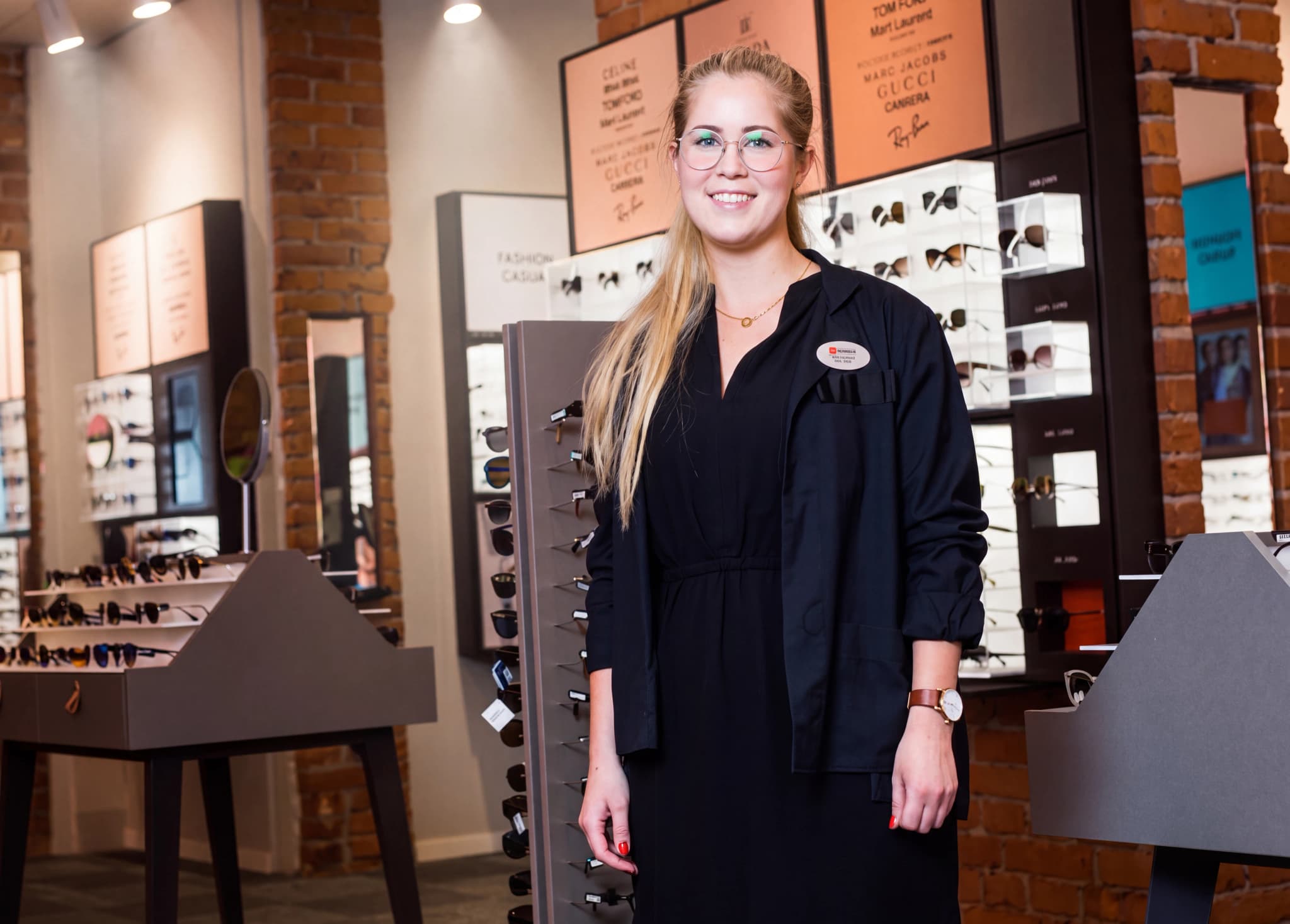 En optiker med glasögon och ett namnskylt på sig står i en optikerbutik omgiven av glasögonställ.