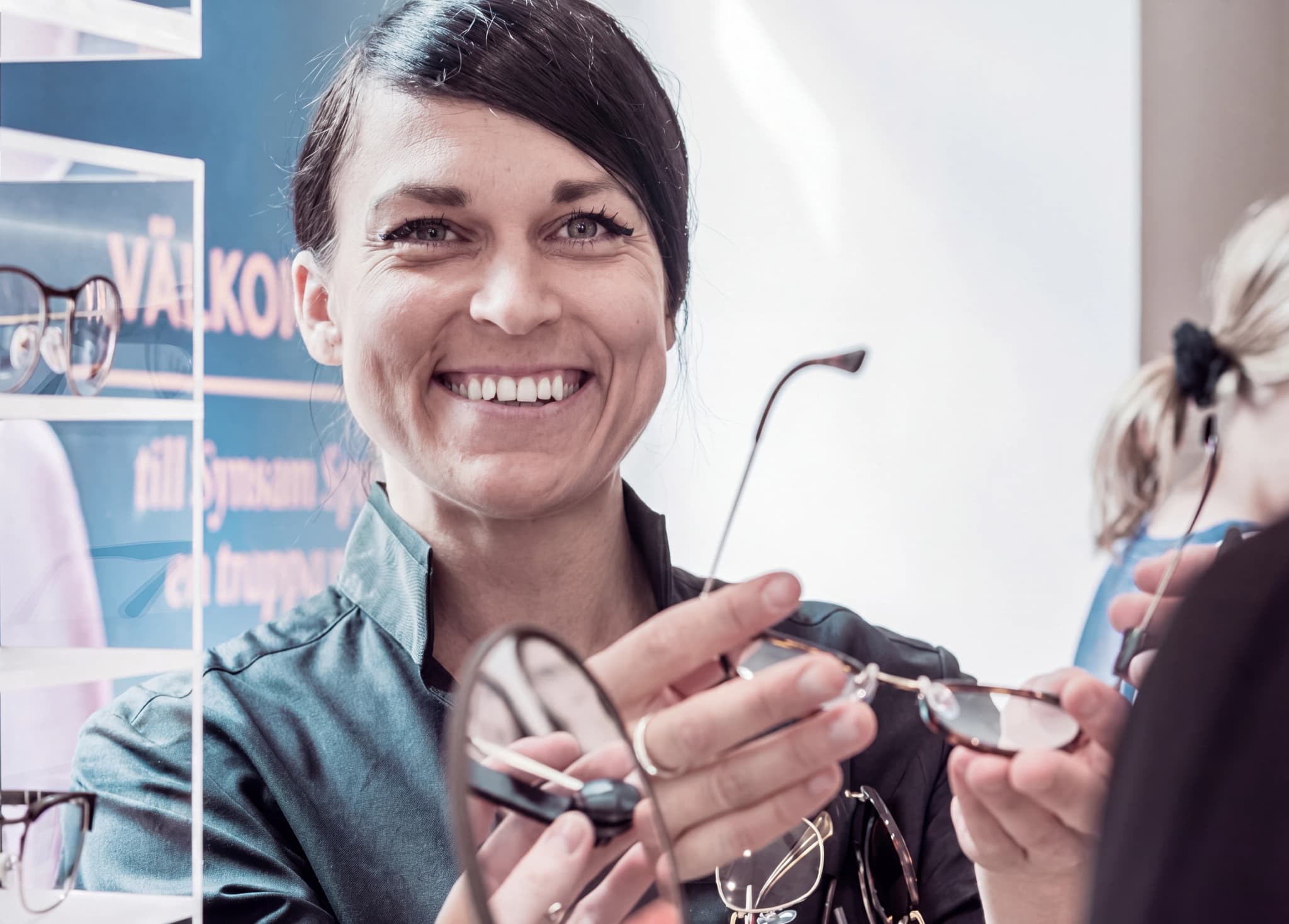 En optiker ler och visar ett par glasögon för en patient i en butik.