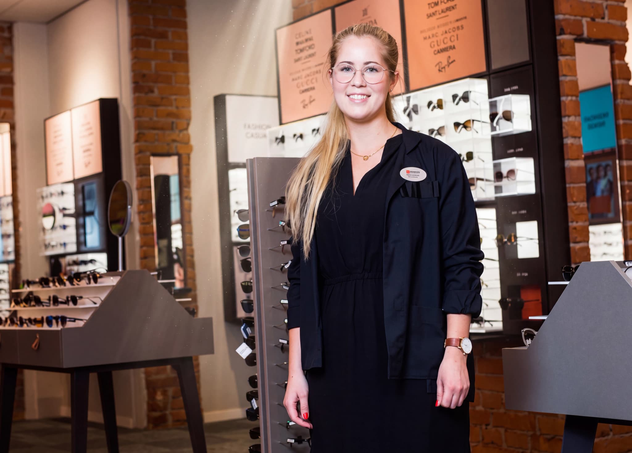 En leende kvinna i glasögon står framför hyllor med glasögon i en butik.