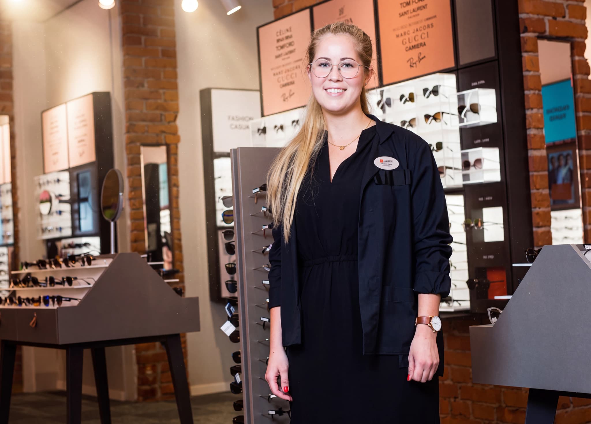 En kvinnlig optiker med glasögon ler vid ett displayställ med glasögon i en butik.