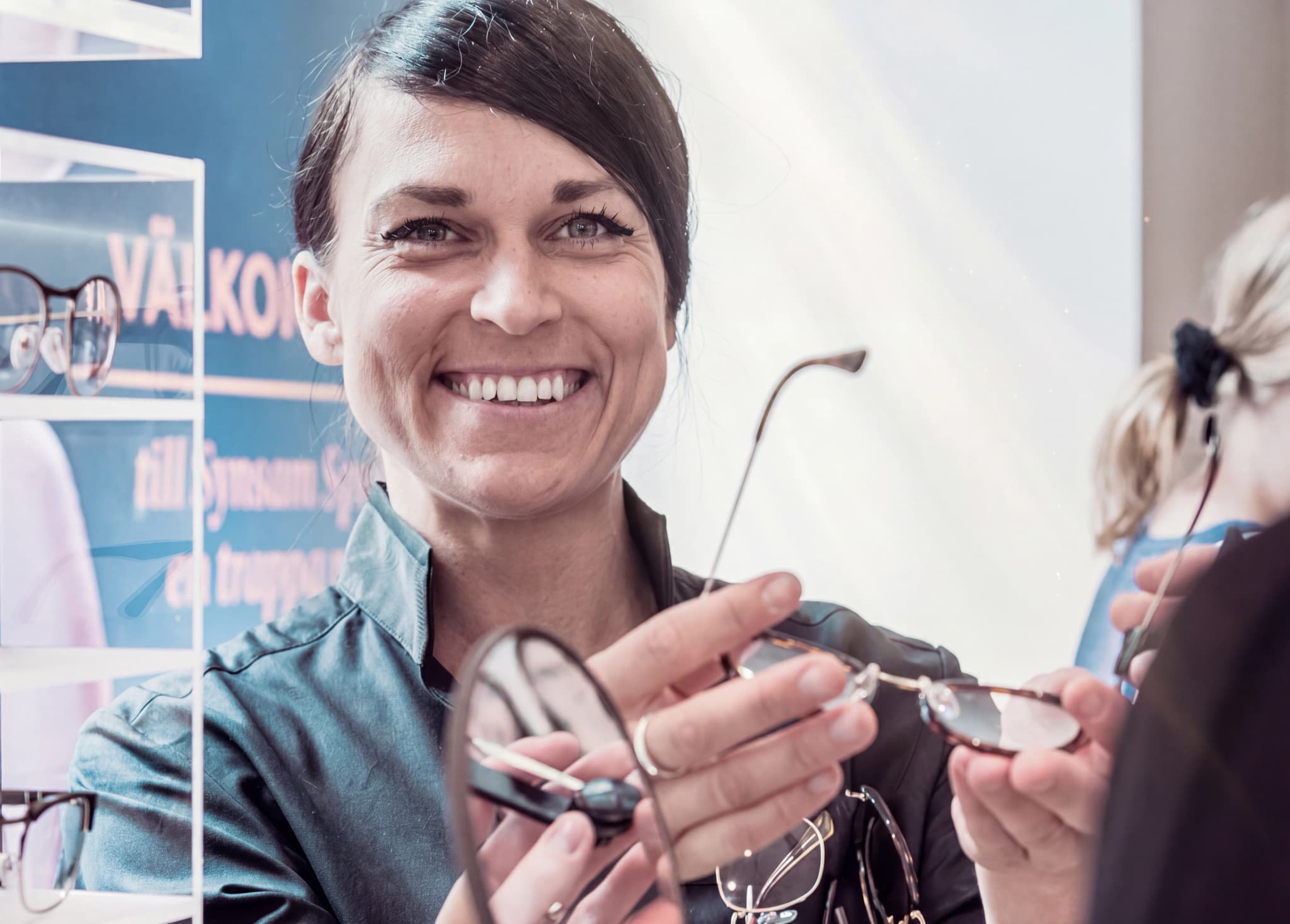 Optiker visar glasögon till kund framför en vägg med glasögon.