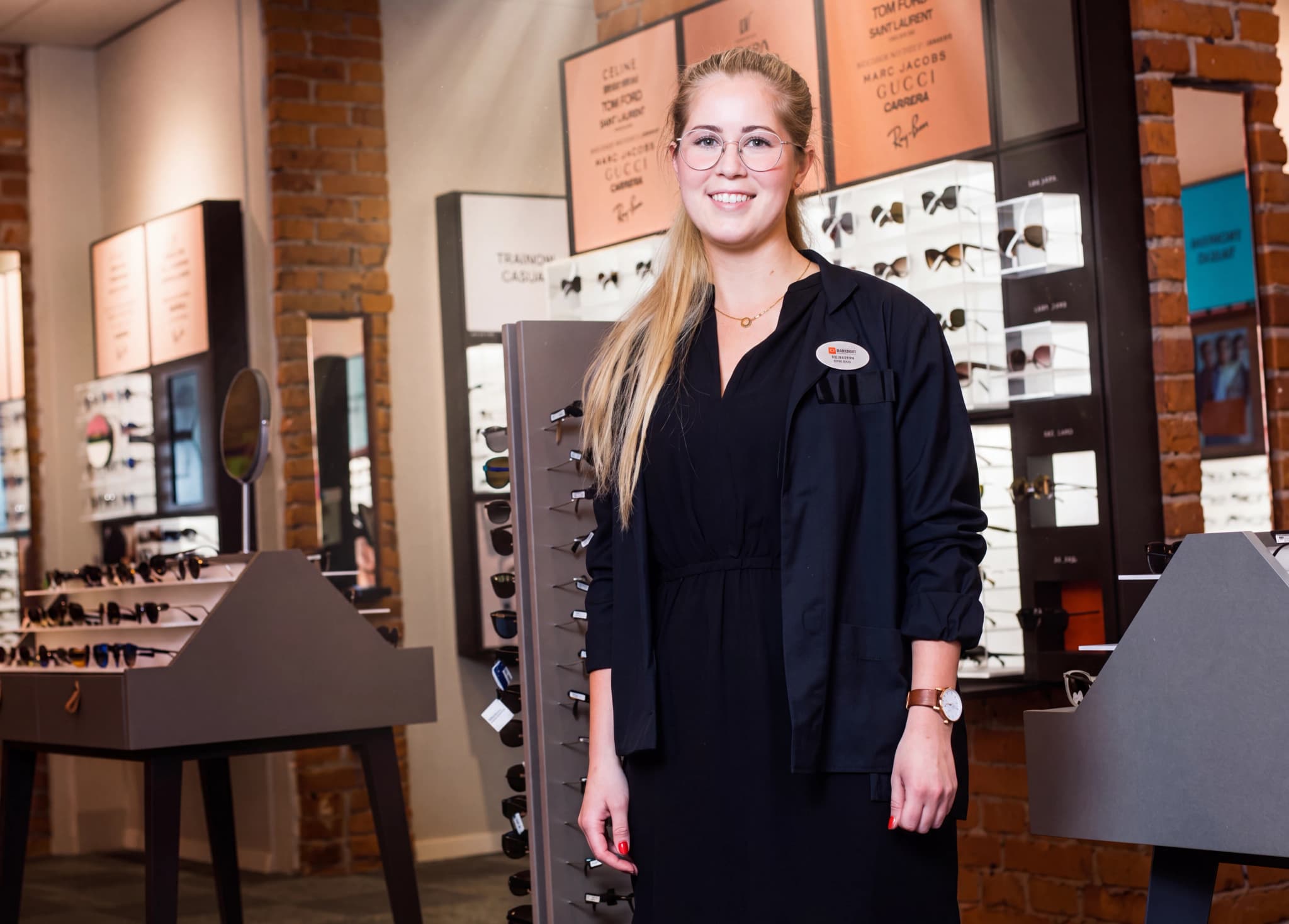 En kvinnlig optiker står i en butik och ler mot kameran, med en display av glasögon i bakgrunden.