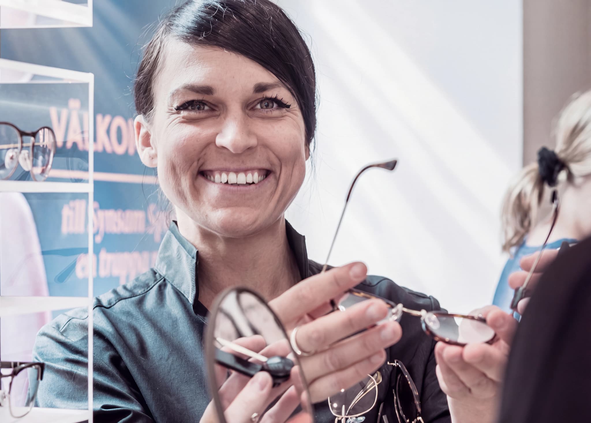 En optiker visar ett par glasögon för en kund i en butik med glasögon på hyllorna i bakgrunden.