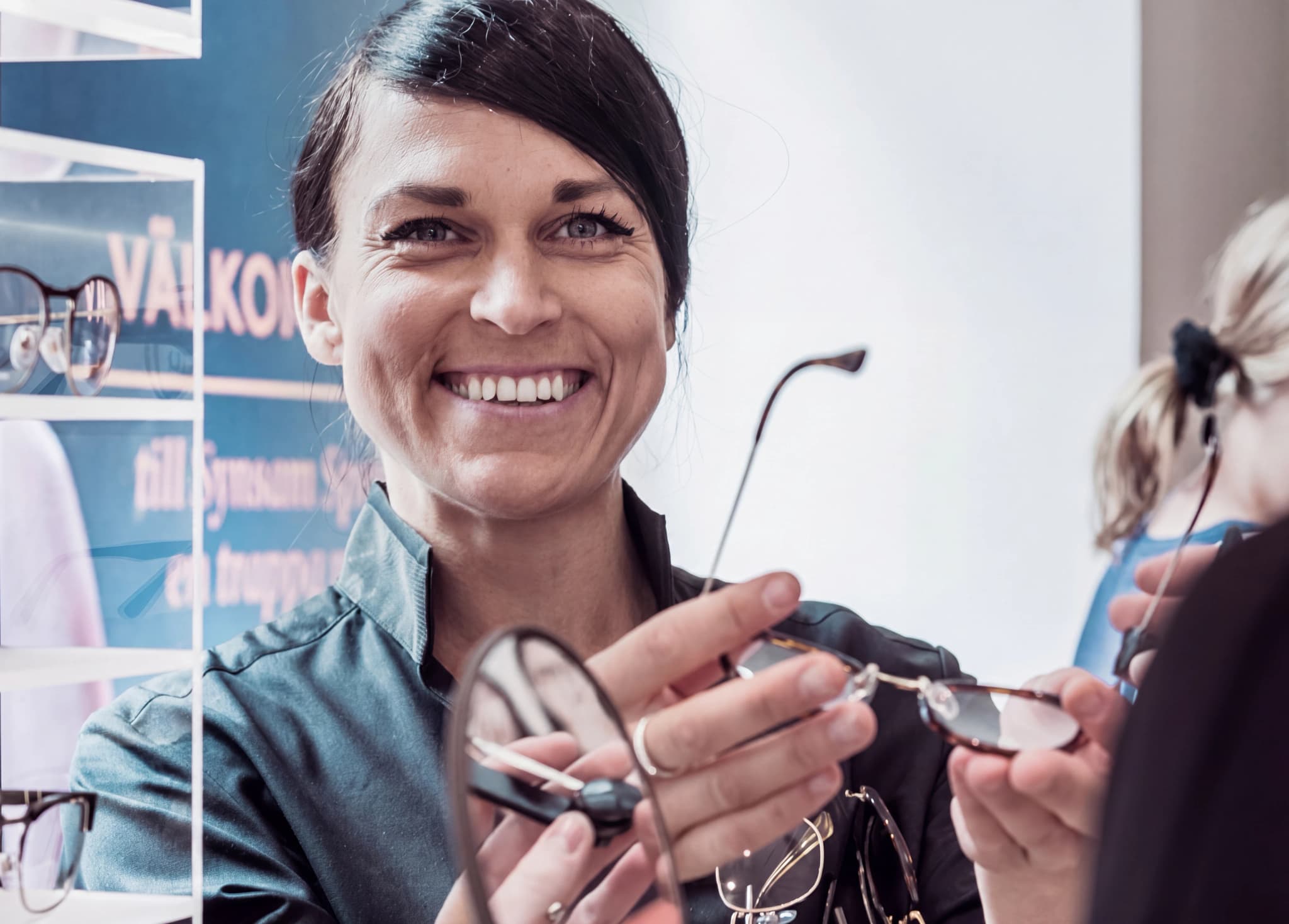 En optiker ler och håller upp ett par glasögon mot en kund i en butik.