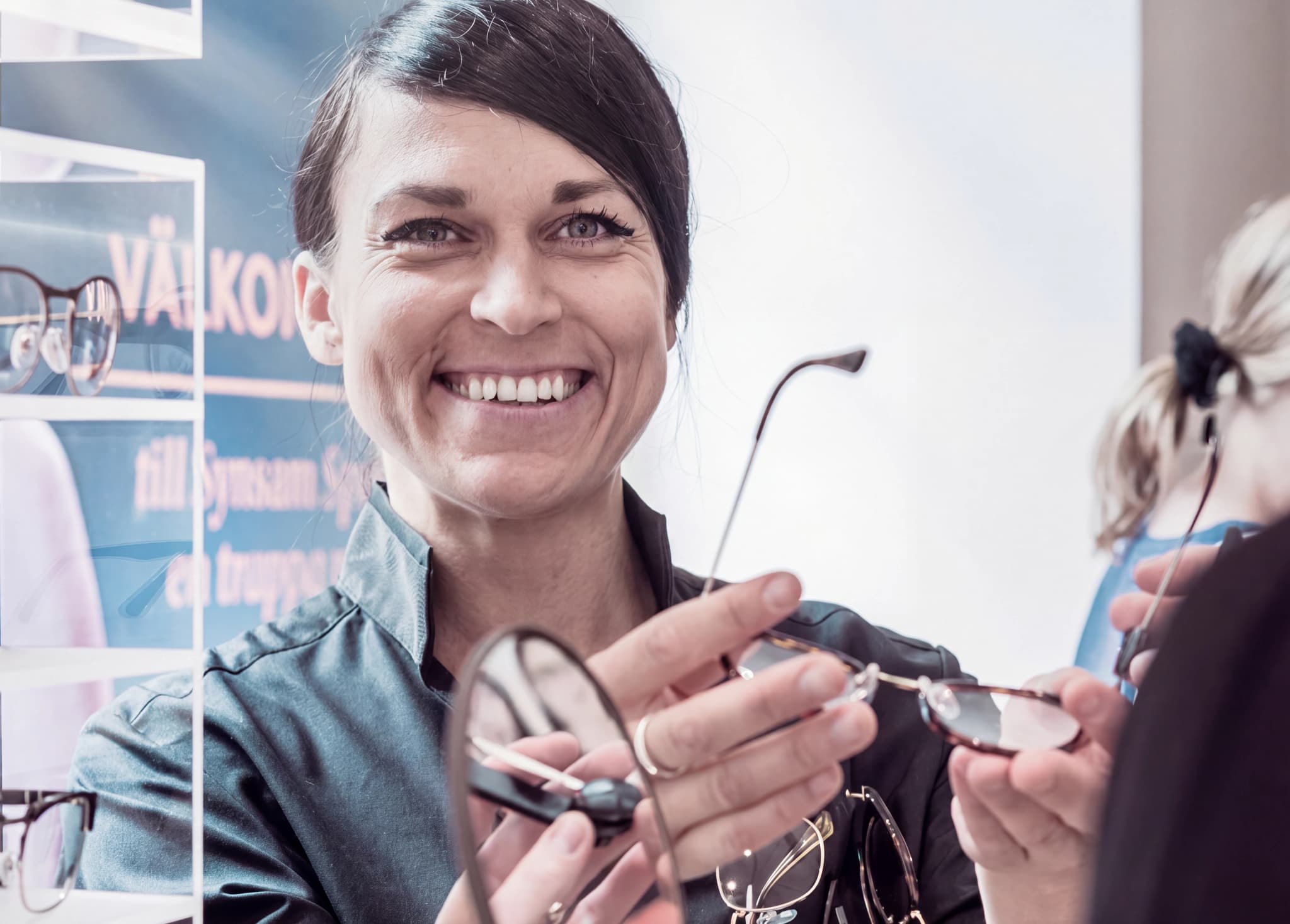 En optiker ler och håller upp ett par glasögon för en kund i en butik.