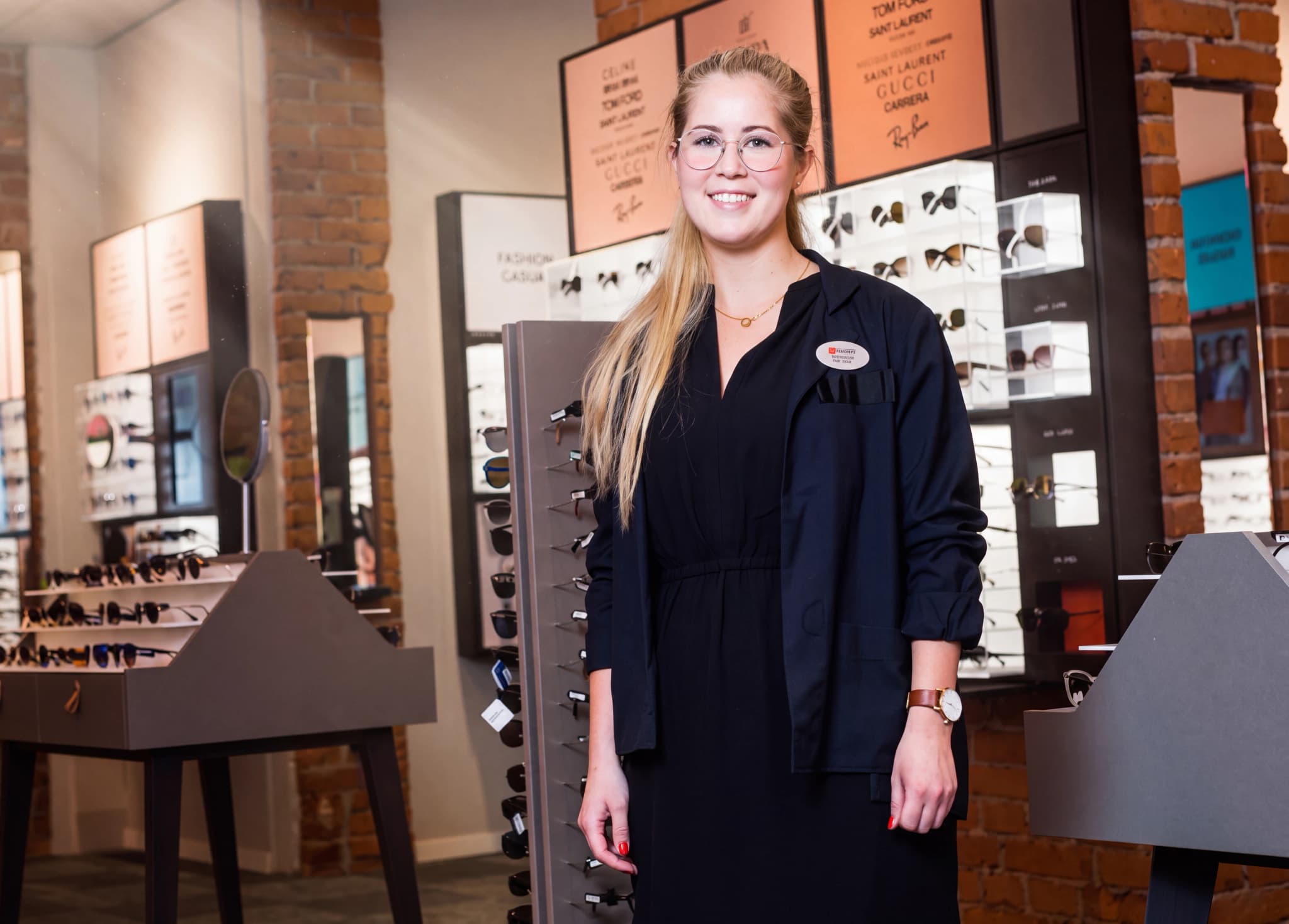 En glad optiker i glasögon och svart klädsel ler mot kameran i en optikerbutik med glasögon displayer.