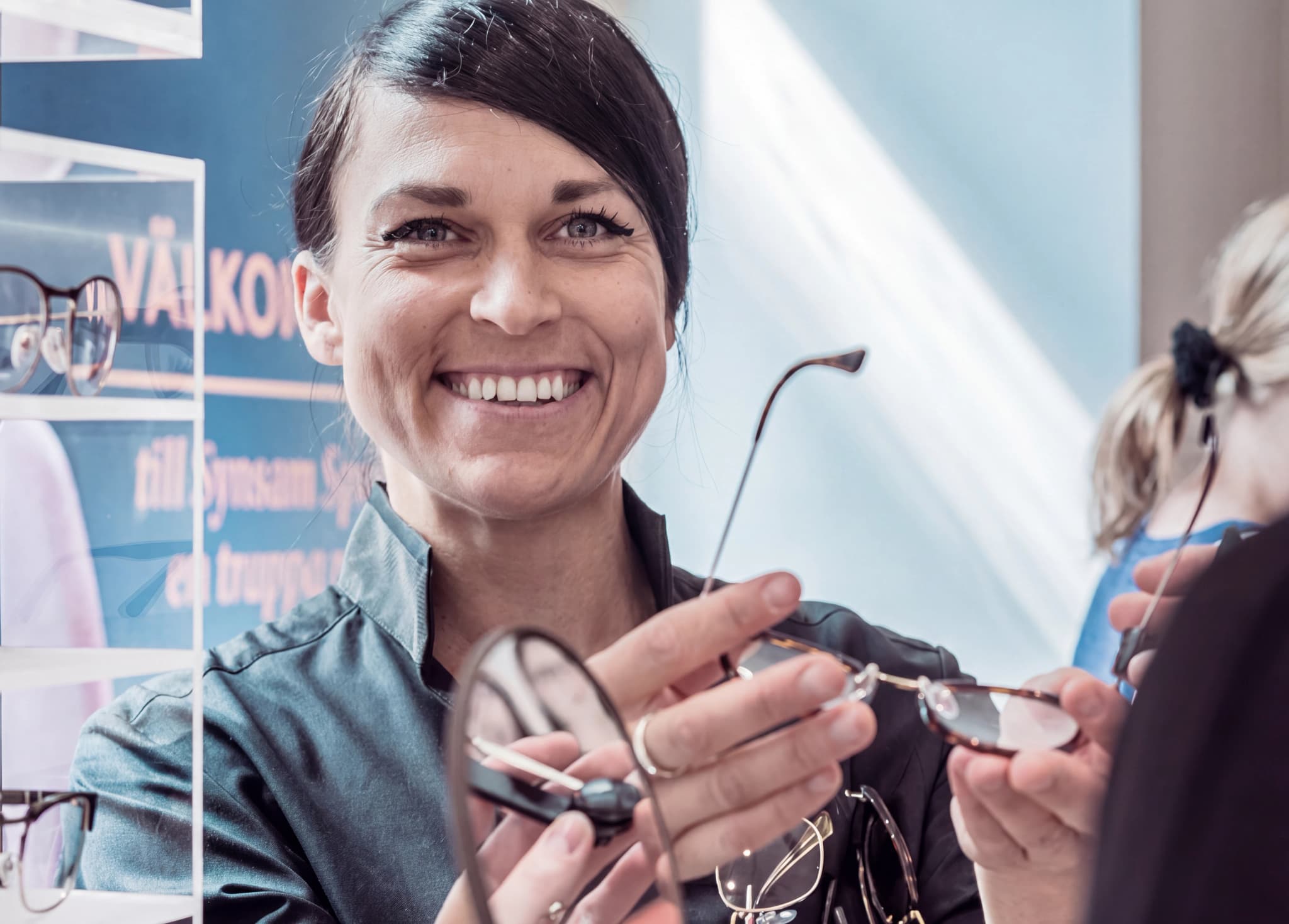 En optiker ler medan hon visar upp ett par glasögon för en kund i en butik.