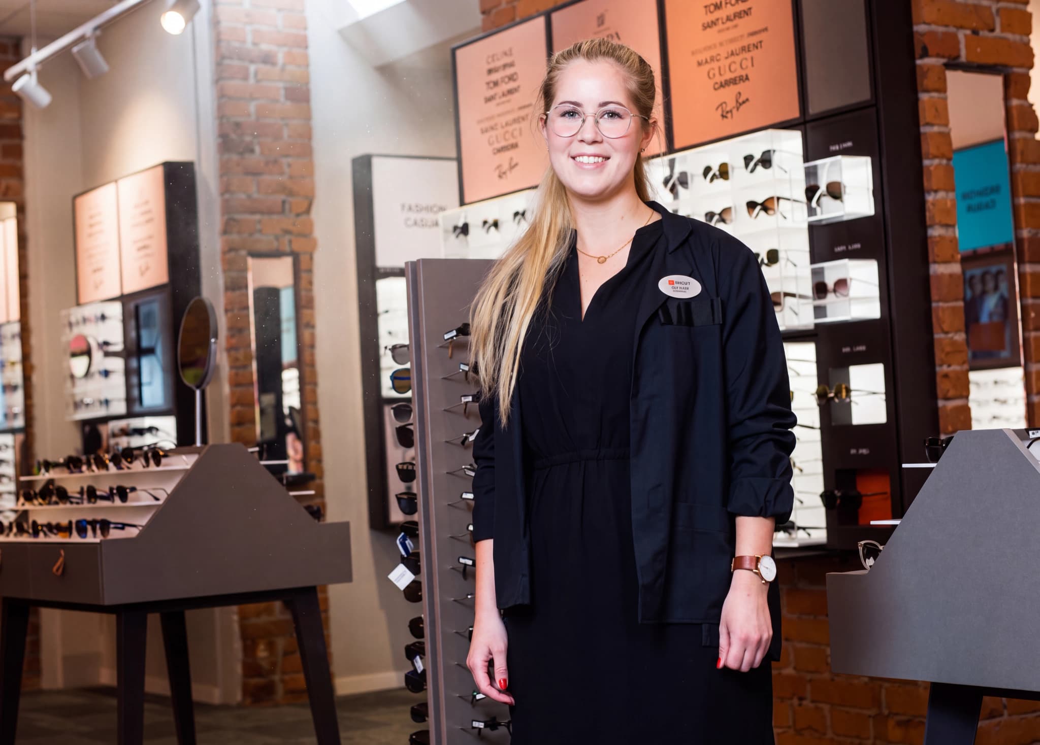 En ung kvinna med glasögon står och ler i en optikerbutik med hyllor fyllda med glasögon bakom sig.