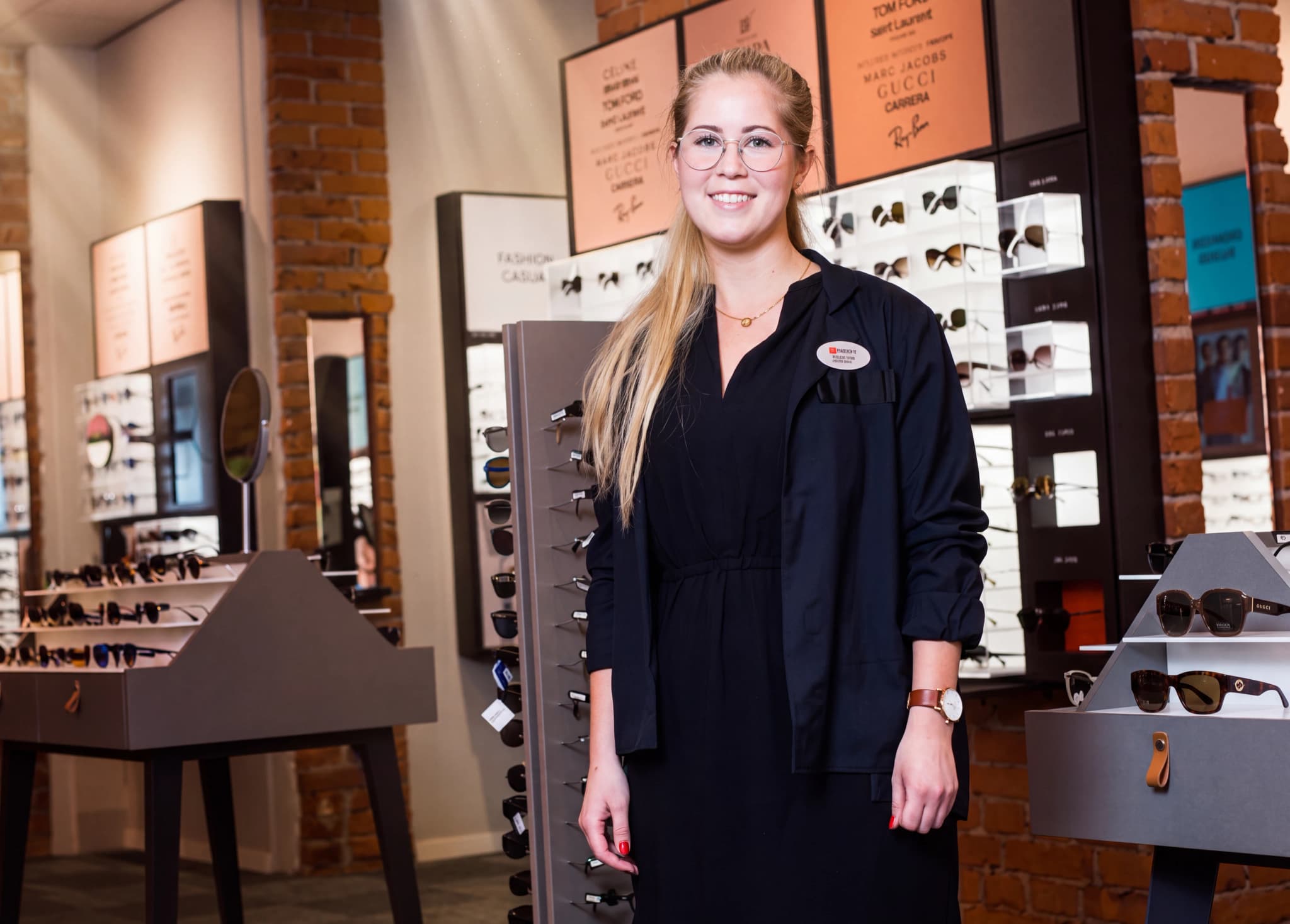 En ung kvinna med glasögon, iklädd en svart skjorta och klänning, står och ler i en optikaffär bredvid en display med glasögon.