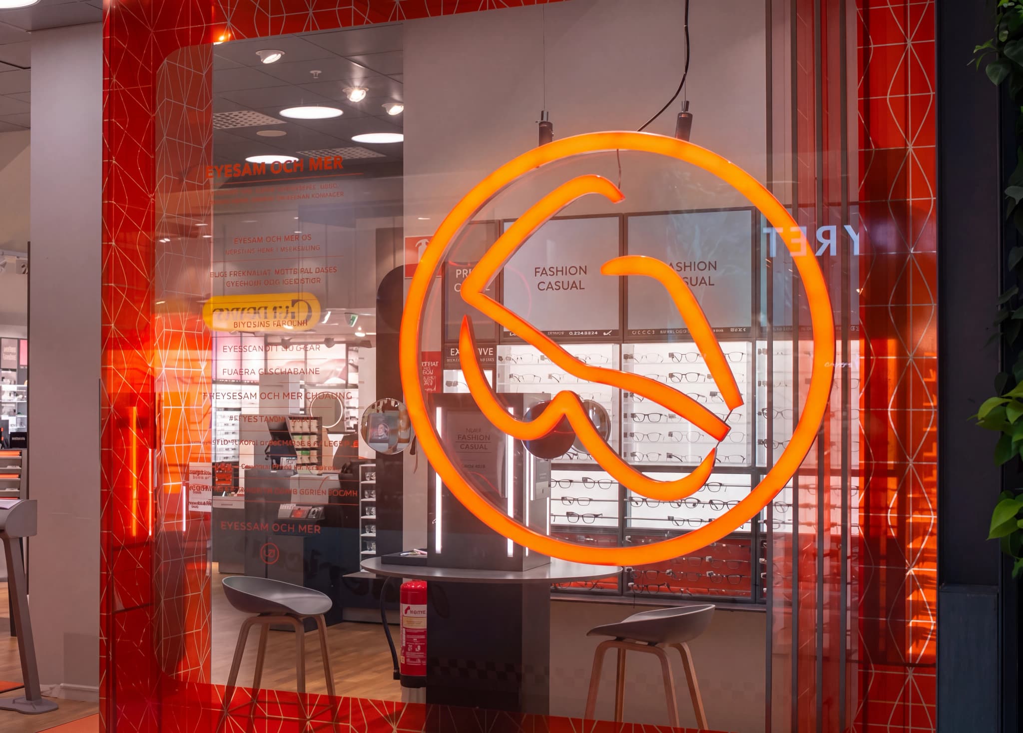 Interiör av en optikerbutik med ett stort lysande orange märke i förgrunden, skyltfönster med glasögon och personal i bakgrunden.