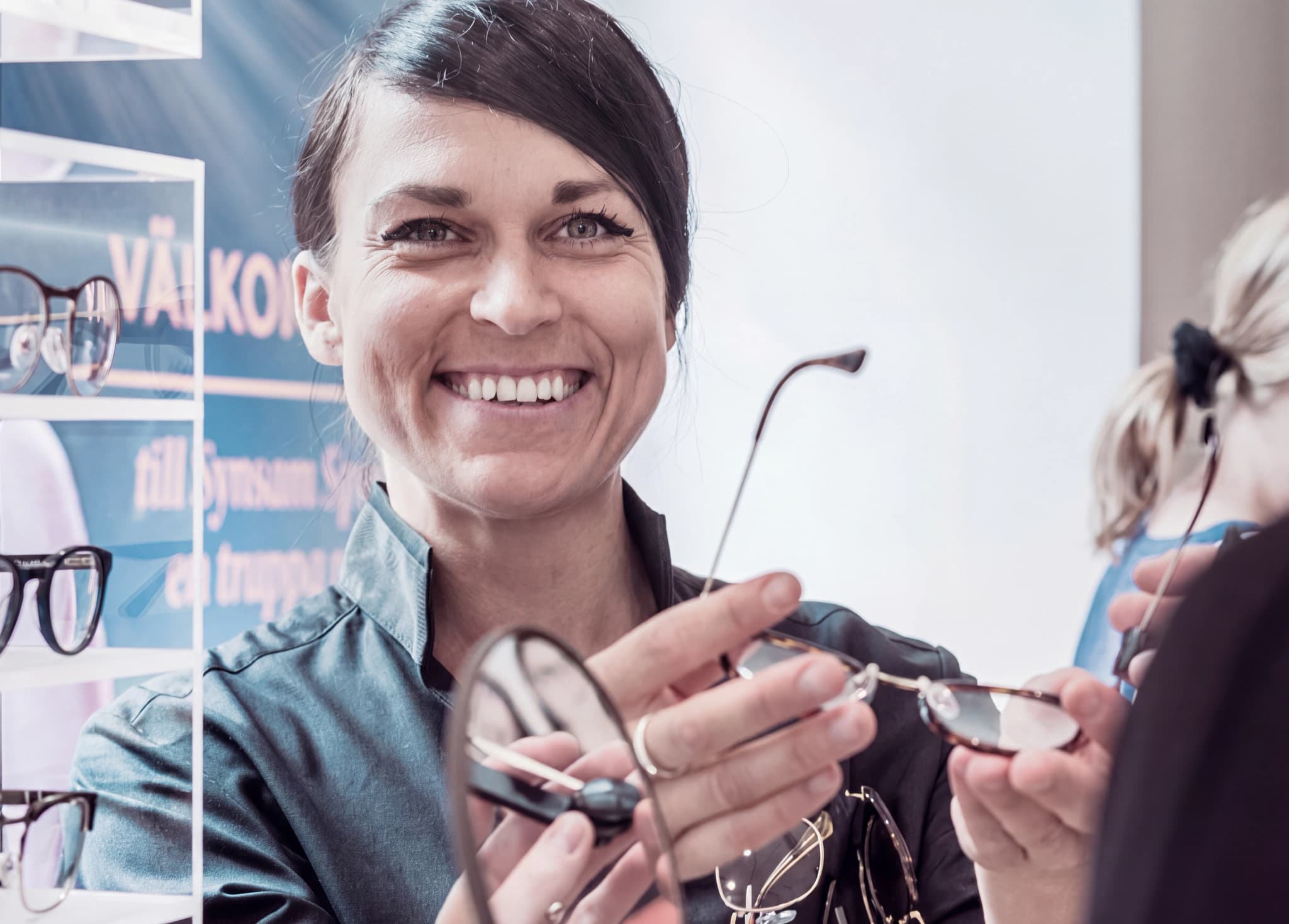 En leende optiker visar ett par glasögon för en kund i en butik med glasögonställningar bakom.