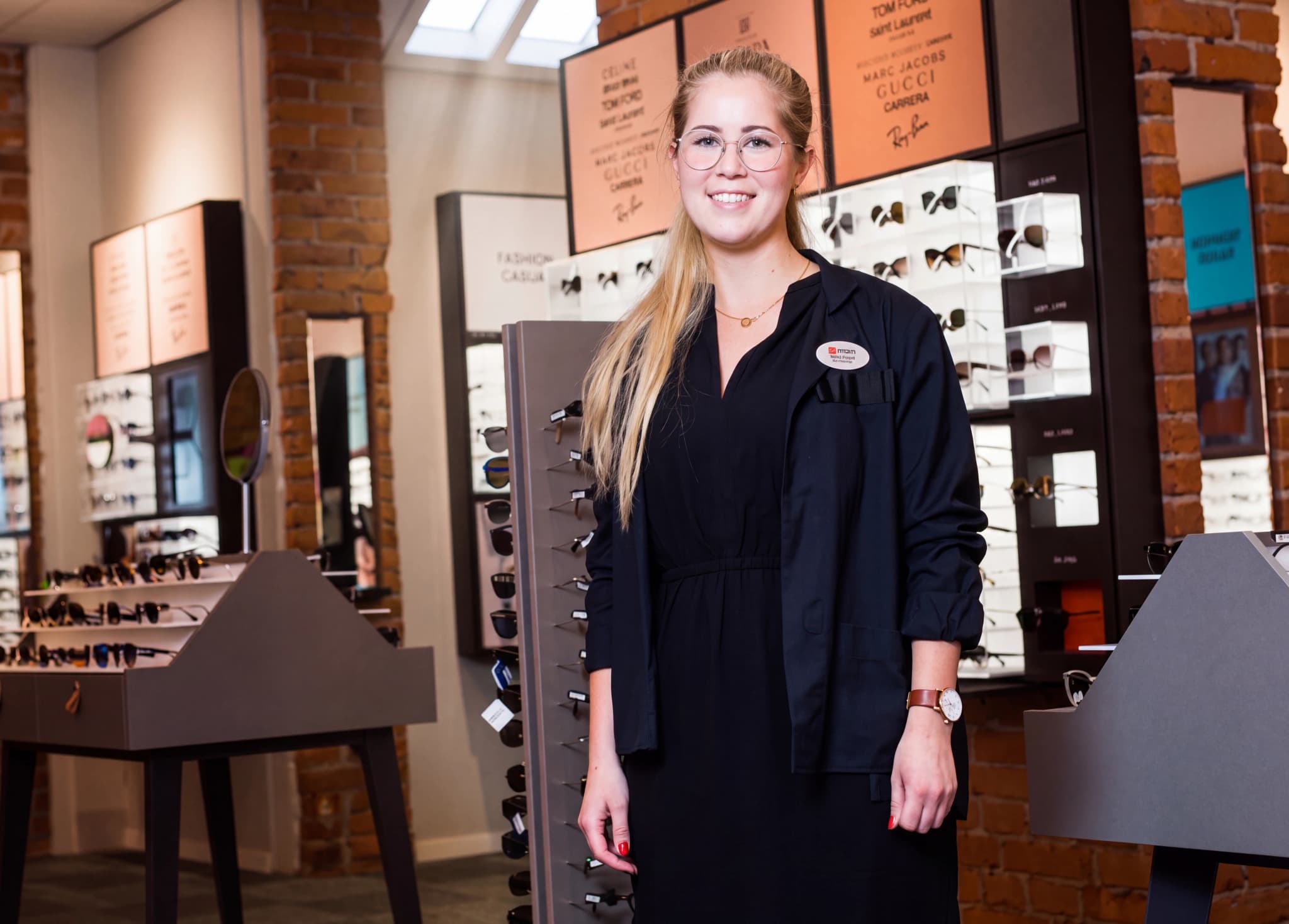 En leende optiker i glasögonbutik står framför hyllor med glasögon och solglasögon.