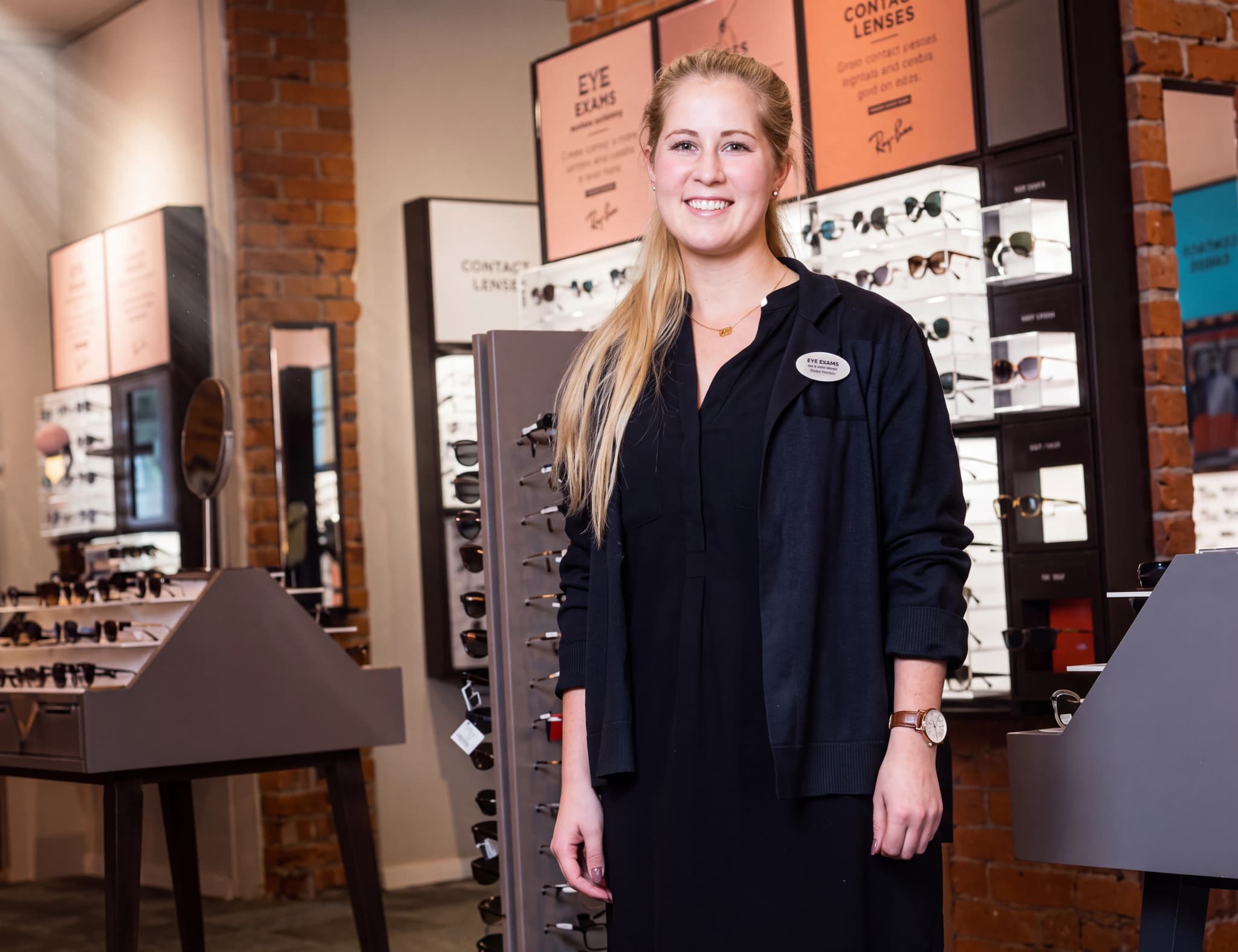Optiker som står i en butik och ler, omgiven av glasögon