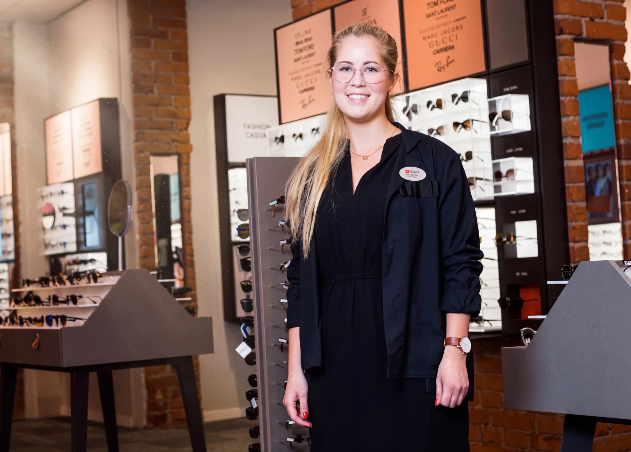En ung kvinna med glasögon och en mörk jacka står i en butik med glasögonhyllor.