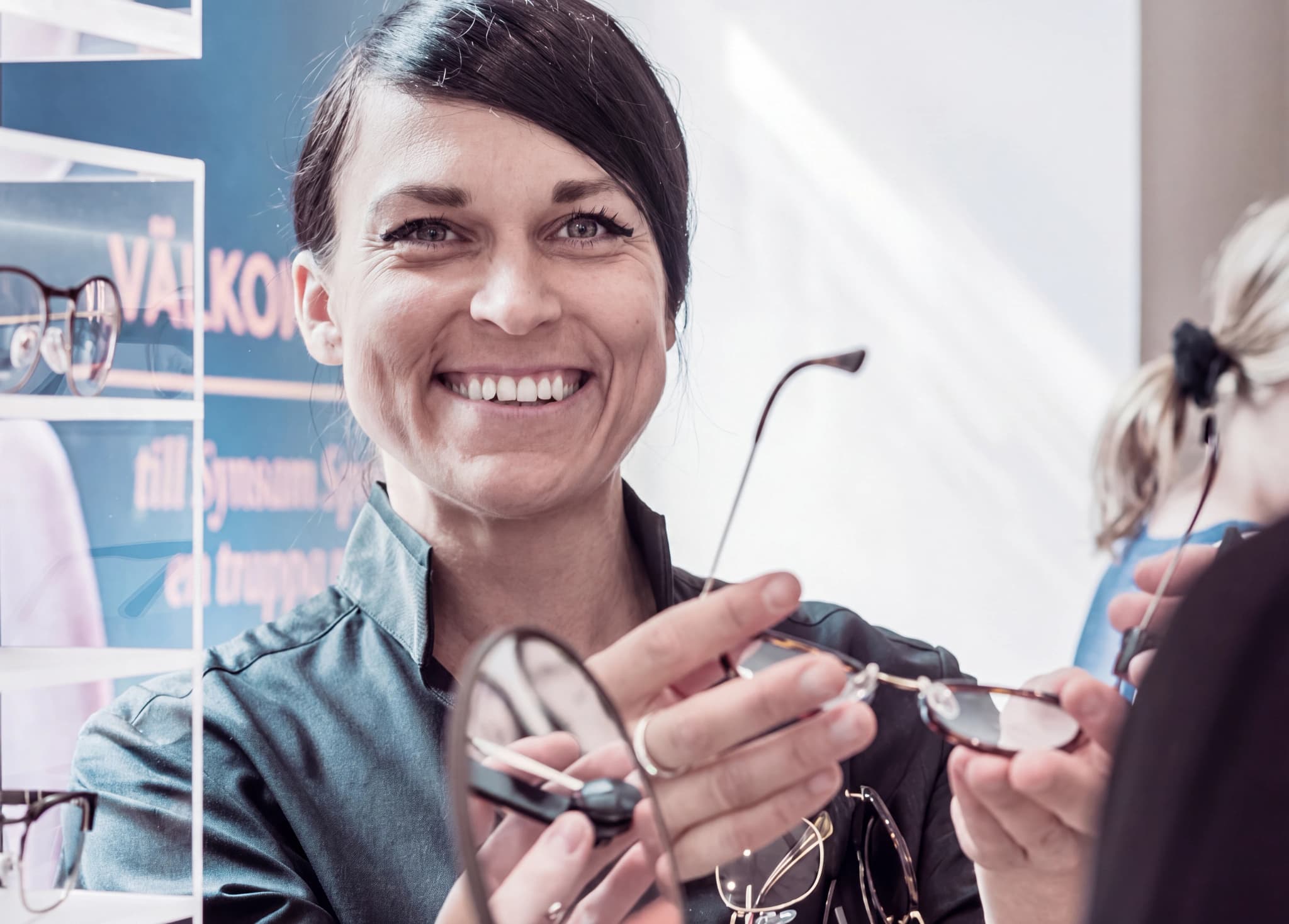 En optiker ler och visar ett par glasögon till en kund i en butik, med andra glasögon synliga i bakgrunden.