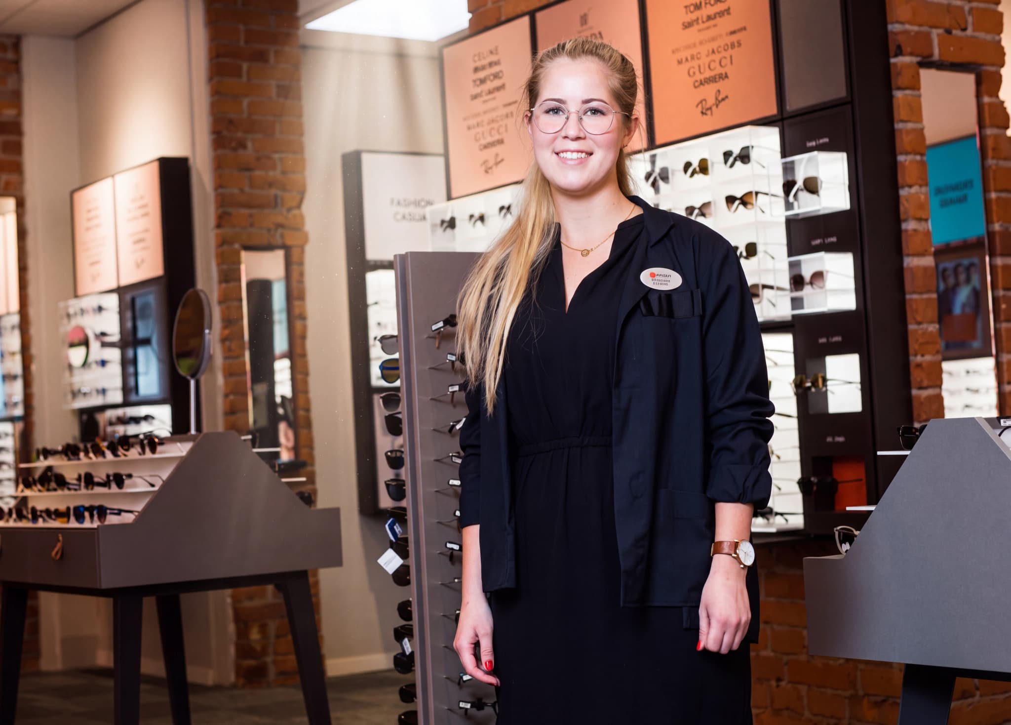 En ung kvinna med glasögon och en svart arbetskläder står i en butik som säljer glasögon.