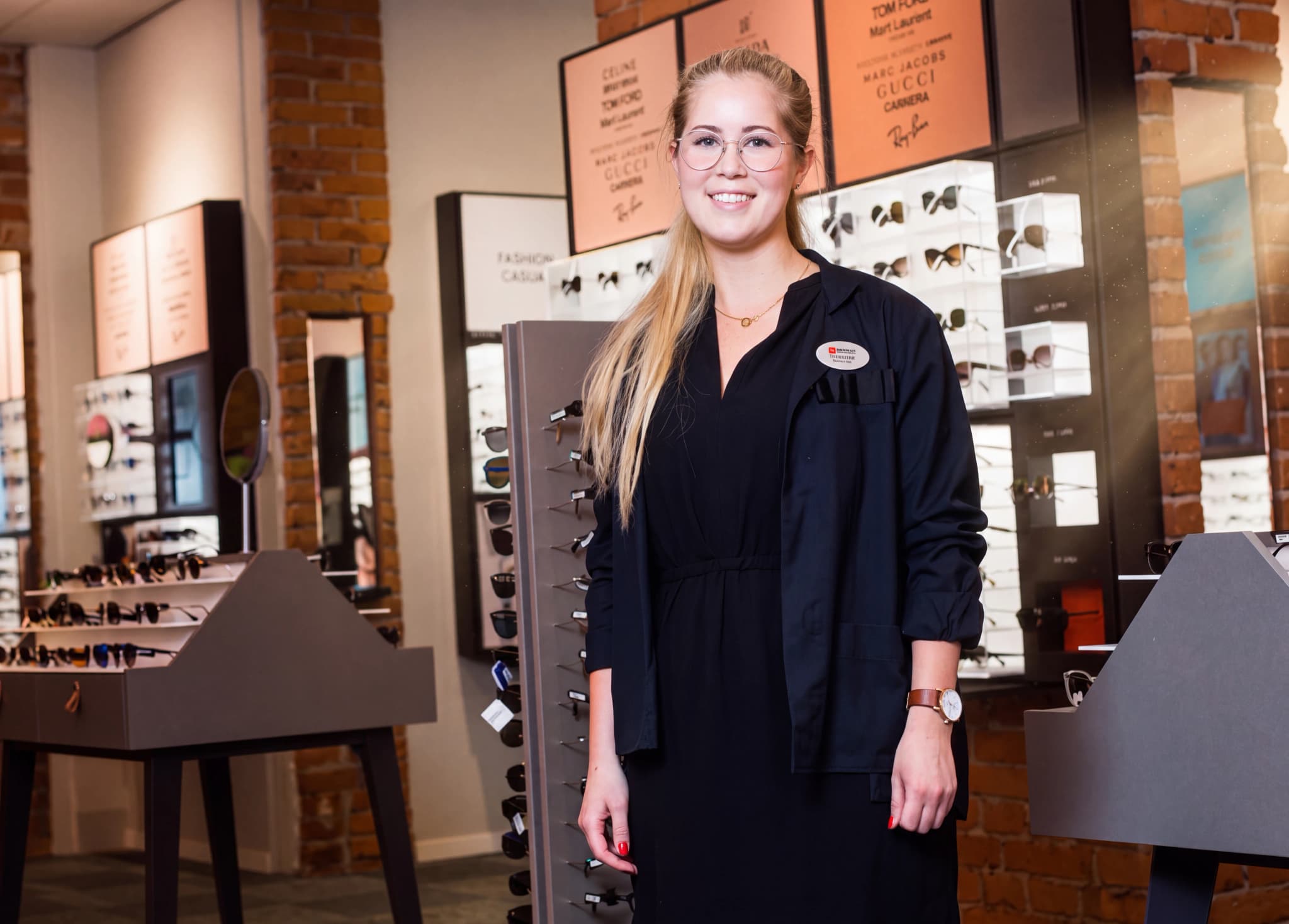 En ung kvinna med glasögon står framför hyllor med glasögon i en butik.