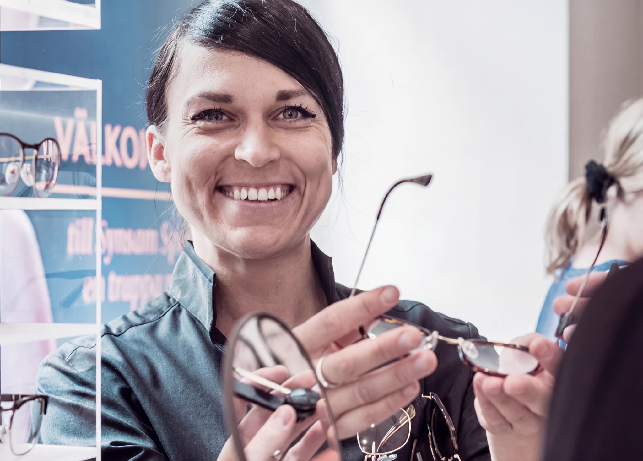Optiker ler och visar ett par glasögon för en kund i en butik med glasögon på hyllor i bakgrunden.