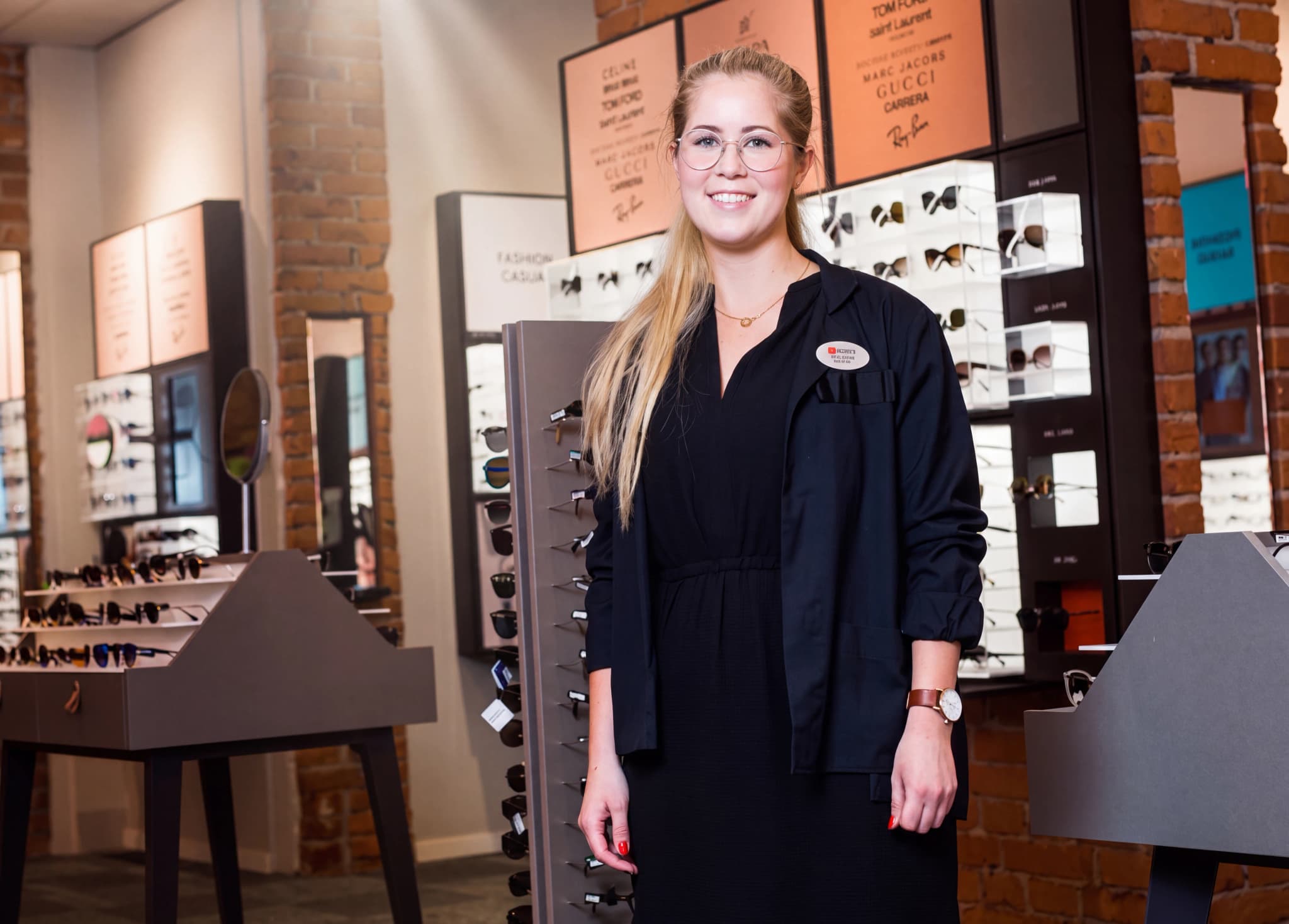 En ung kvinna med glasögon, klädd i en svart överdel, står framför en hylla med glasögon i en optikerbutik och ler mot kameran.