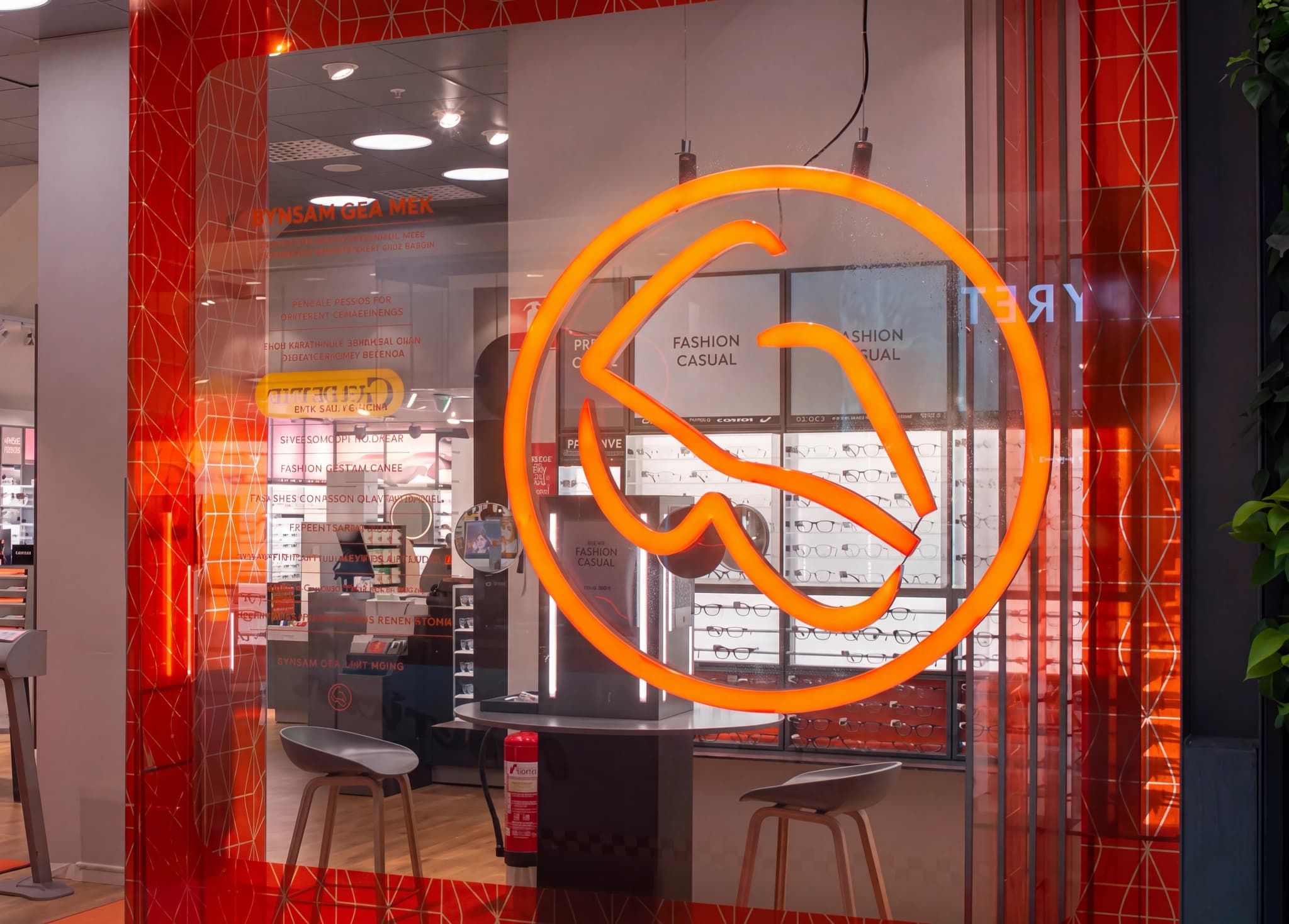 Entrén till en optikerbutik med en stor, lysande orange logotyp framför. Butiken visar upp glasögon och har skyltar för 'FASHION CASUAL'.