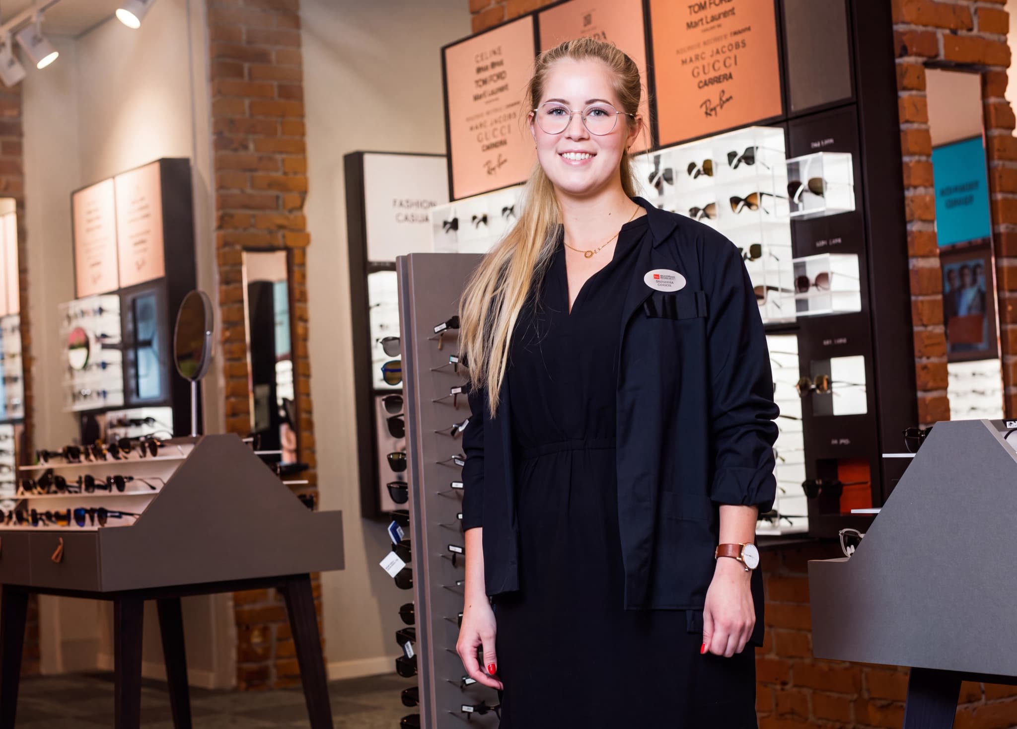 En ung kvinna med glasögon, klädd i en mörkblå arbetsuniform, ler och tittar framåt medan hon står i en butik som säljer glasögon.
