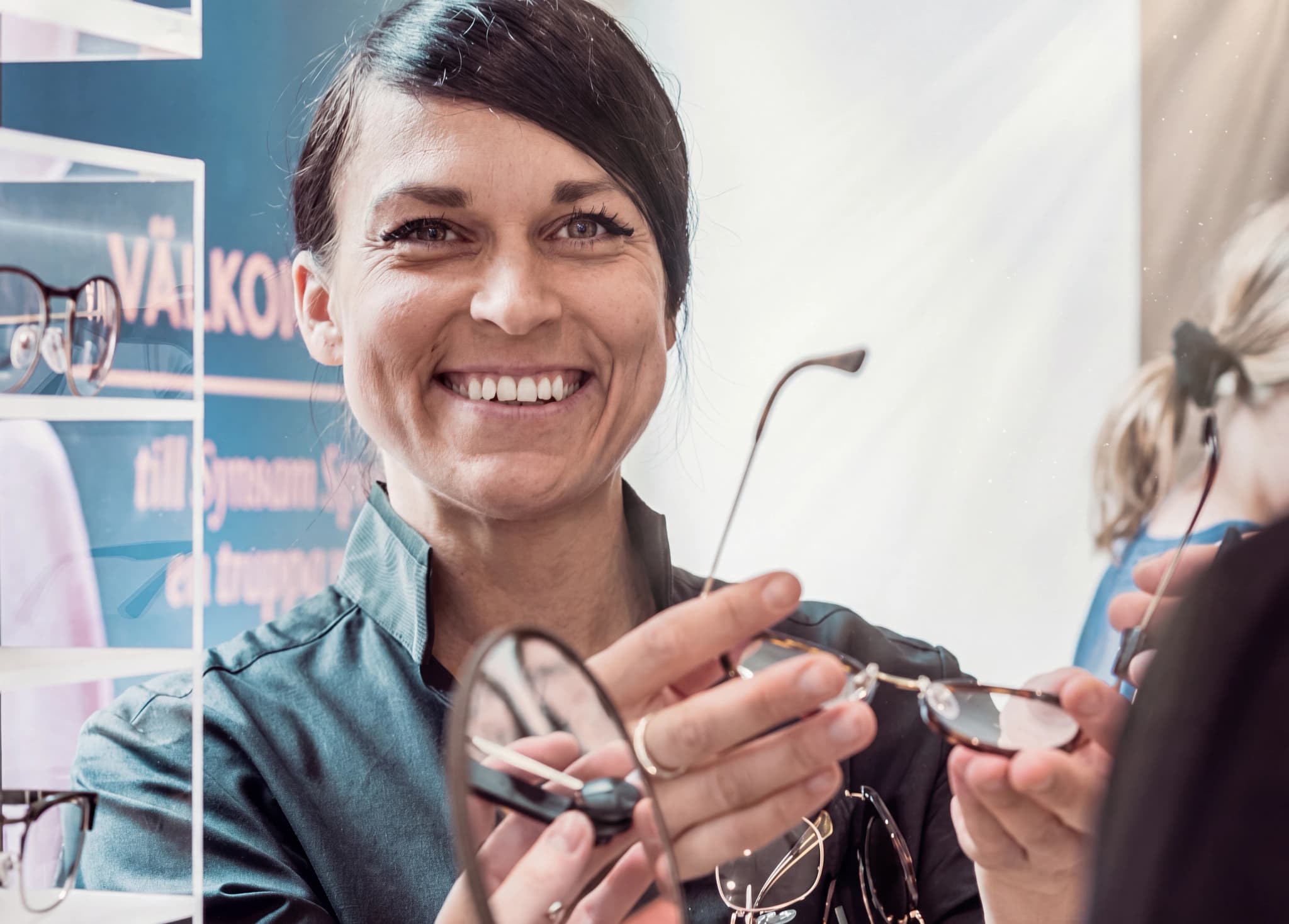 Optiker som ler och håller upp ett par glasögon för en kund i en butik eller mottagning.