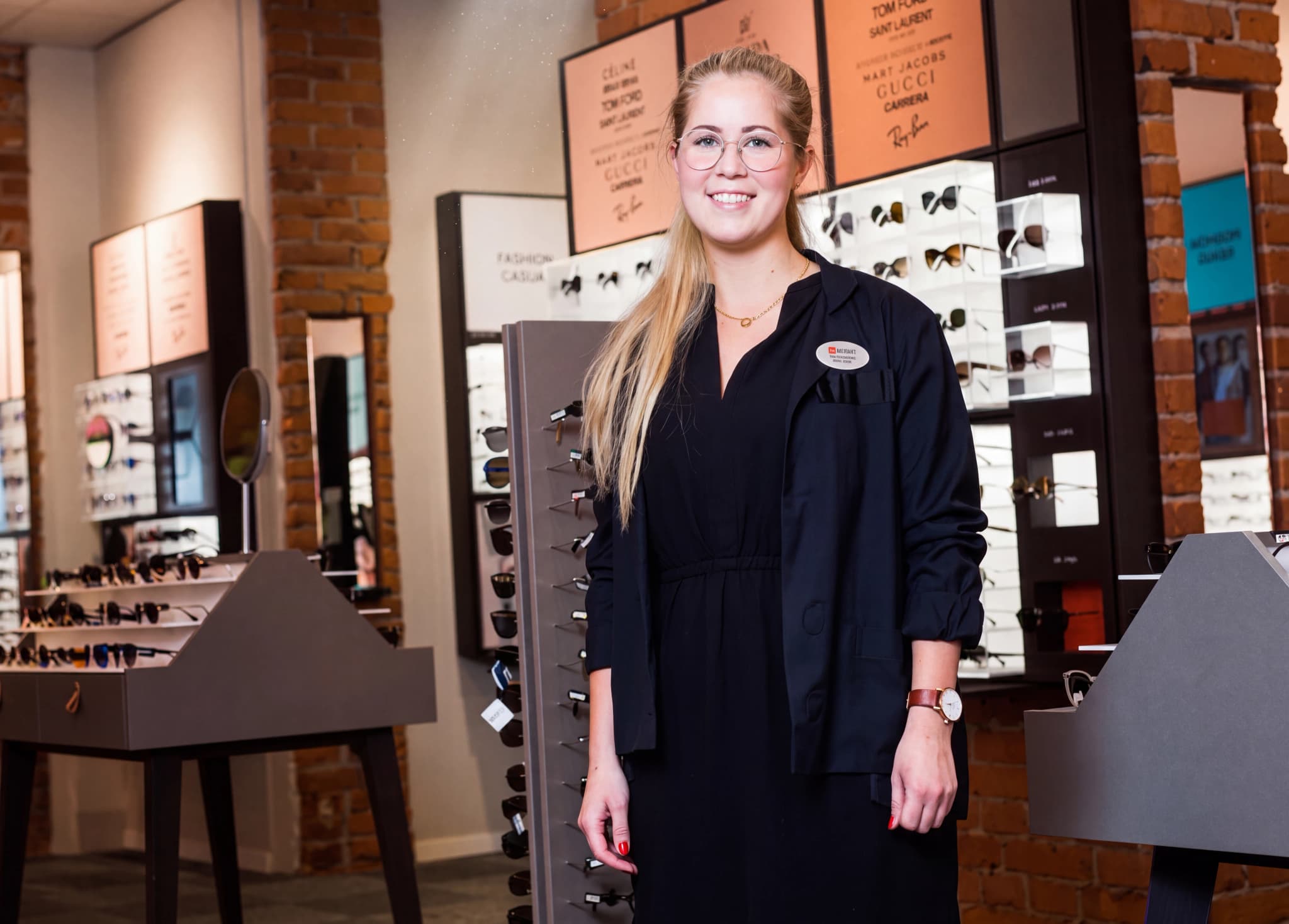 En leende optiker med glasögon med glasögonställningar i bakgrunden.