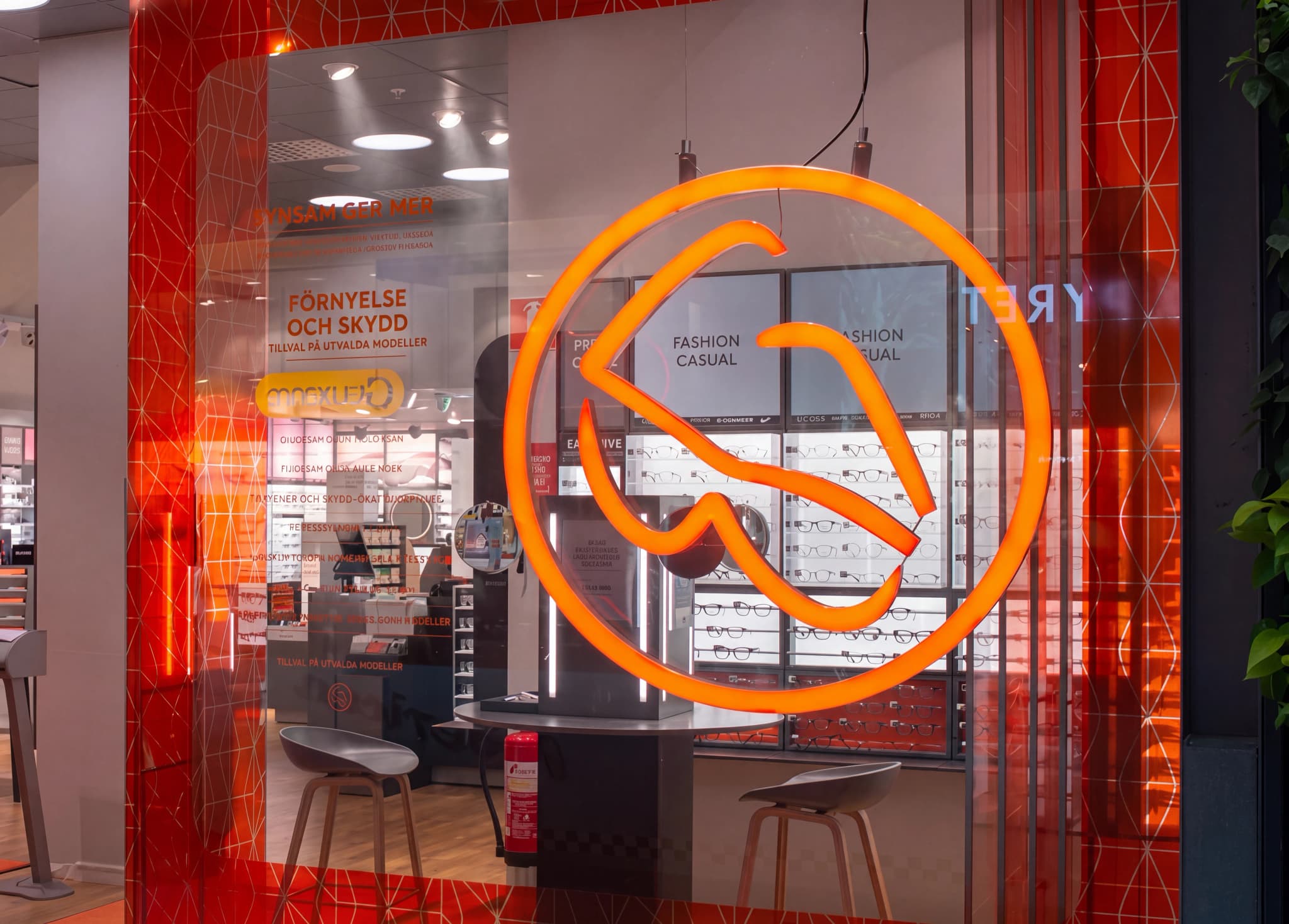 Fönsterdisplay för en optikerbutik med en stor lysande orange logotyp framför glasögonhyllor och skyltar med texten 'FASHION CASUAL'.