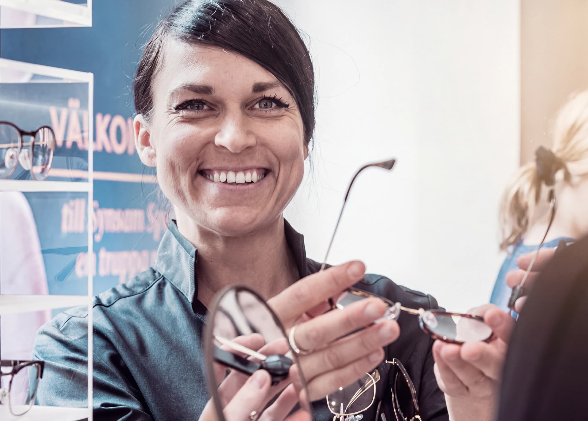 En optiker ler och visar upp ett par glasögon för en kund i en butik.