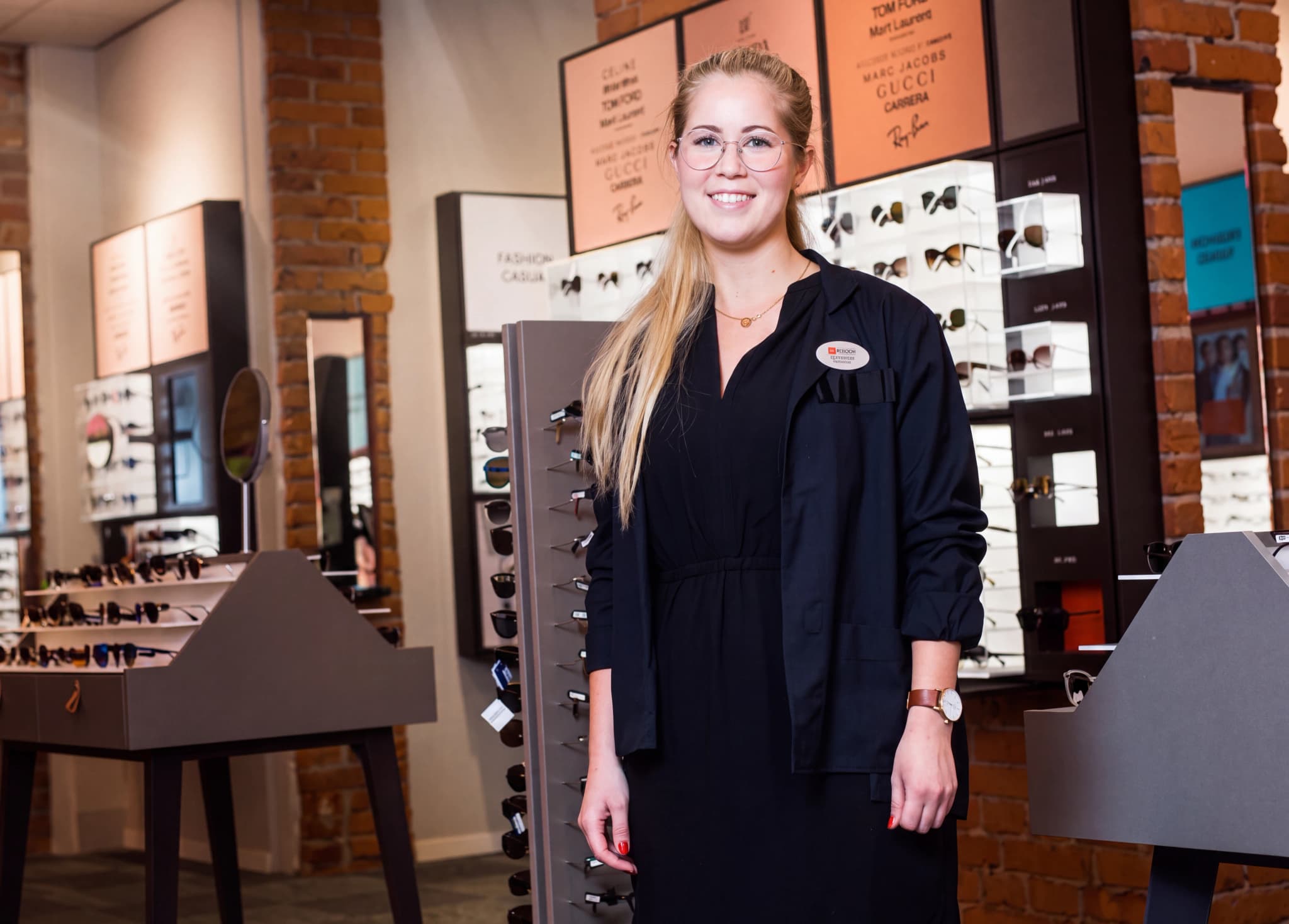 En leende optiker står i en butik som säljer glasögon, med glasögonställningar i bakgrunden.