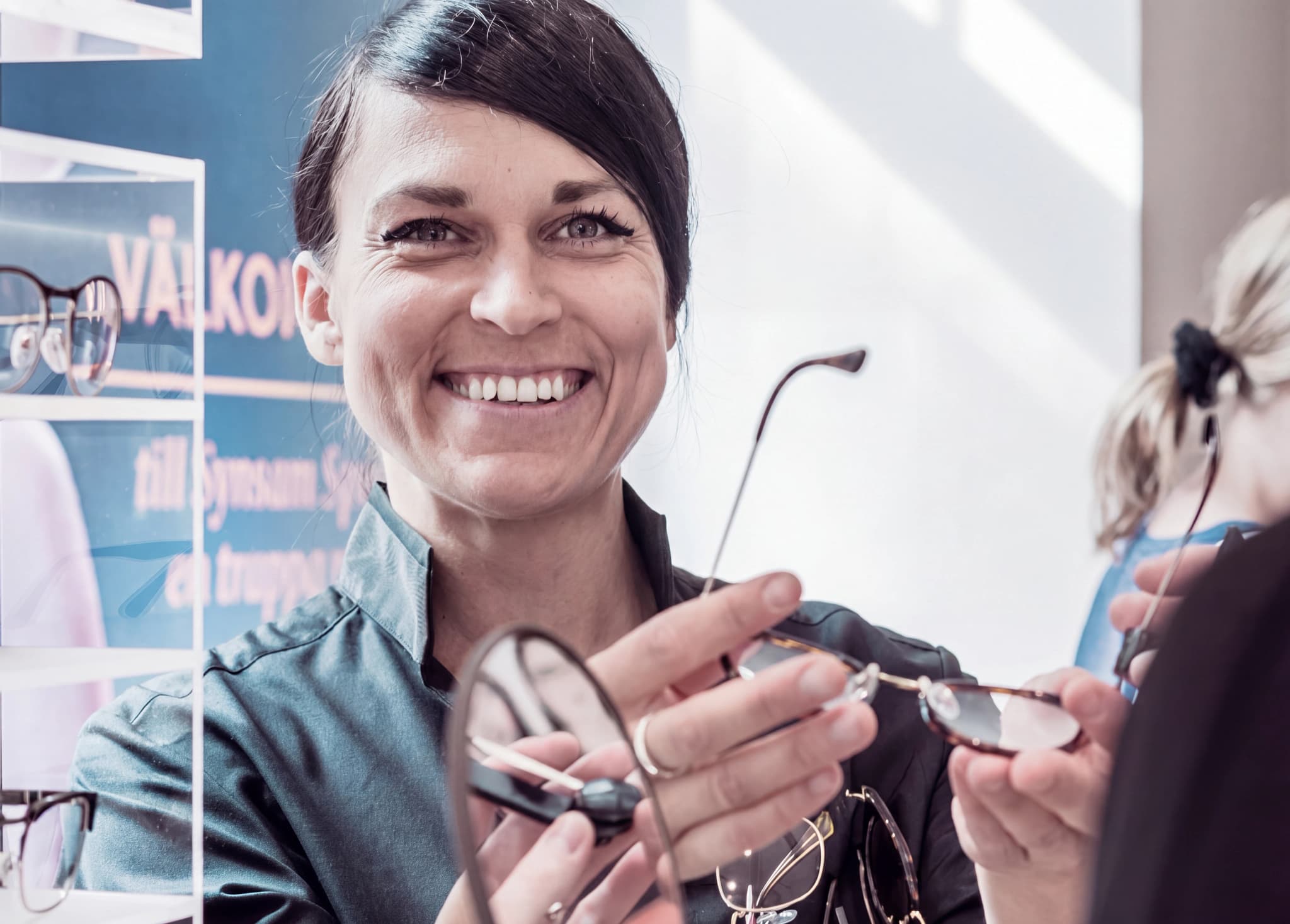 Optiker ler och visar upp ett par glasögon för en kund i en butik.
