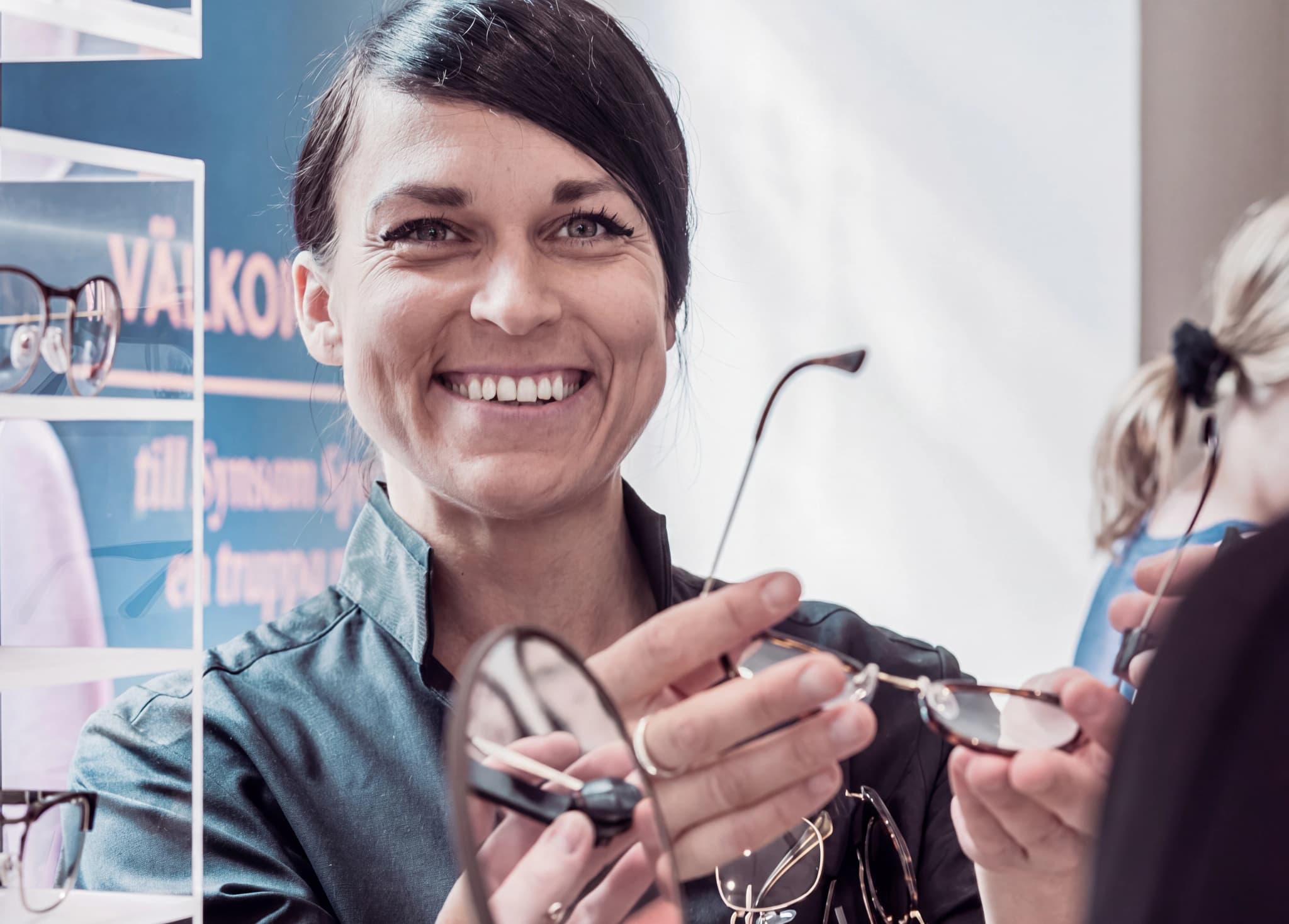 En optiker ler och visar ett par glasögon till en kund i en butik.