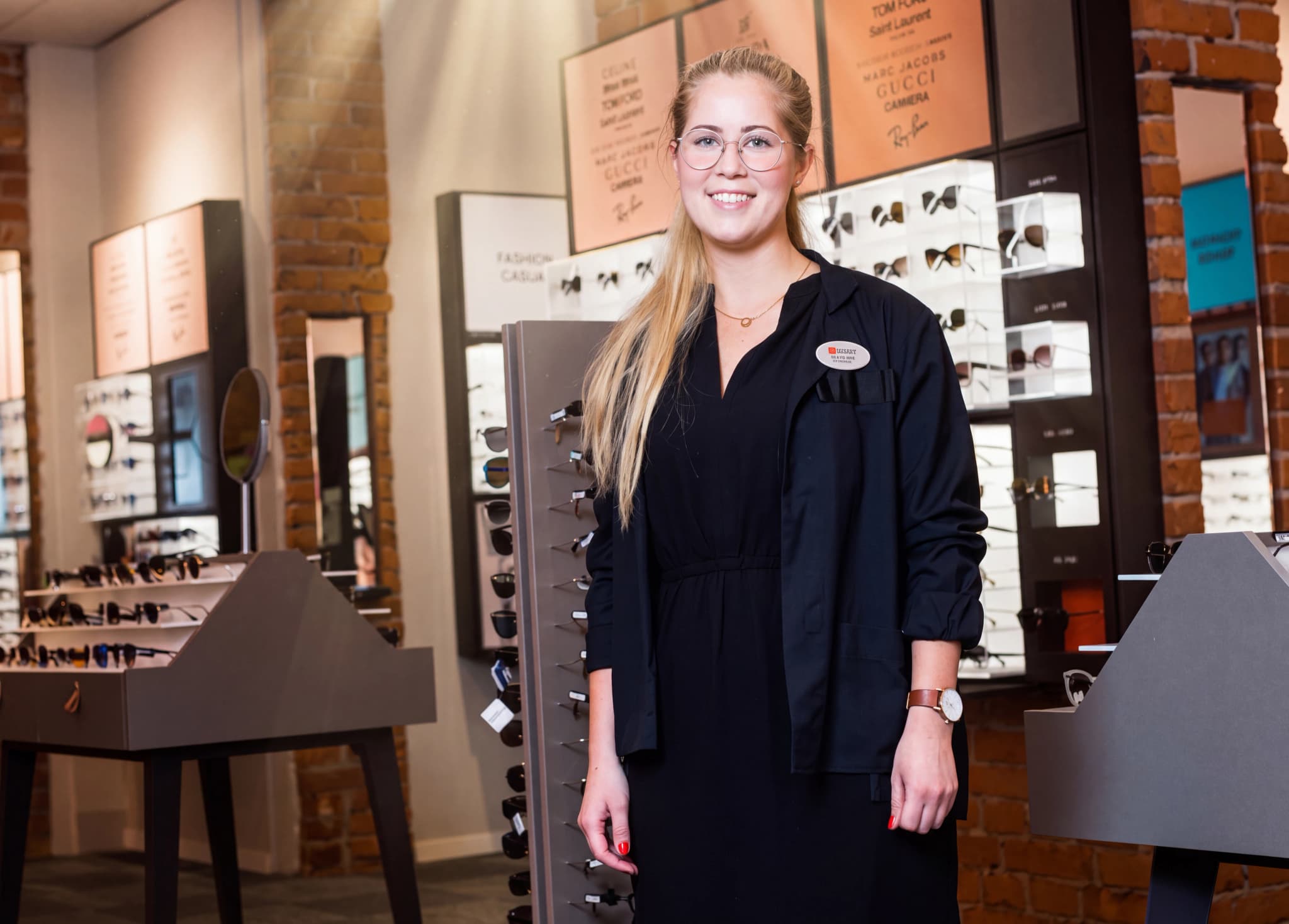 En leende ung kvinna, klädd i en mörk uniform med en namnskylt, står i en butik som säljer glasögon.