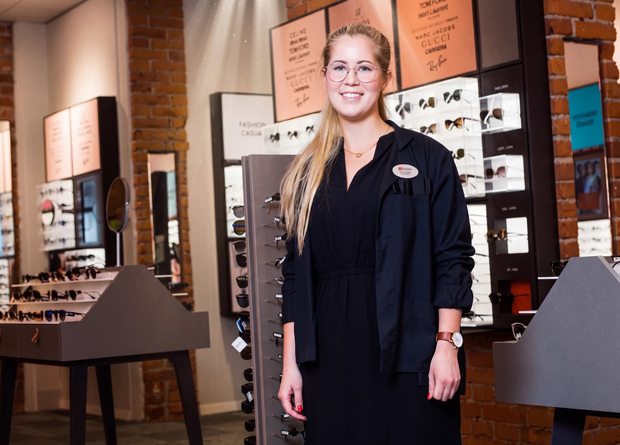 En ung kvinna med glasögon och en svart jacka står och ler i en optikbutik, omgiven av displayer med glasögon.