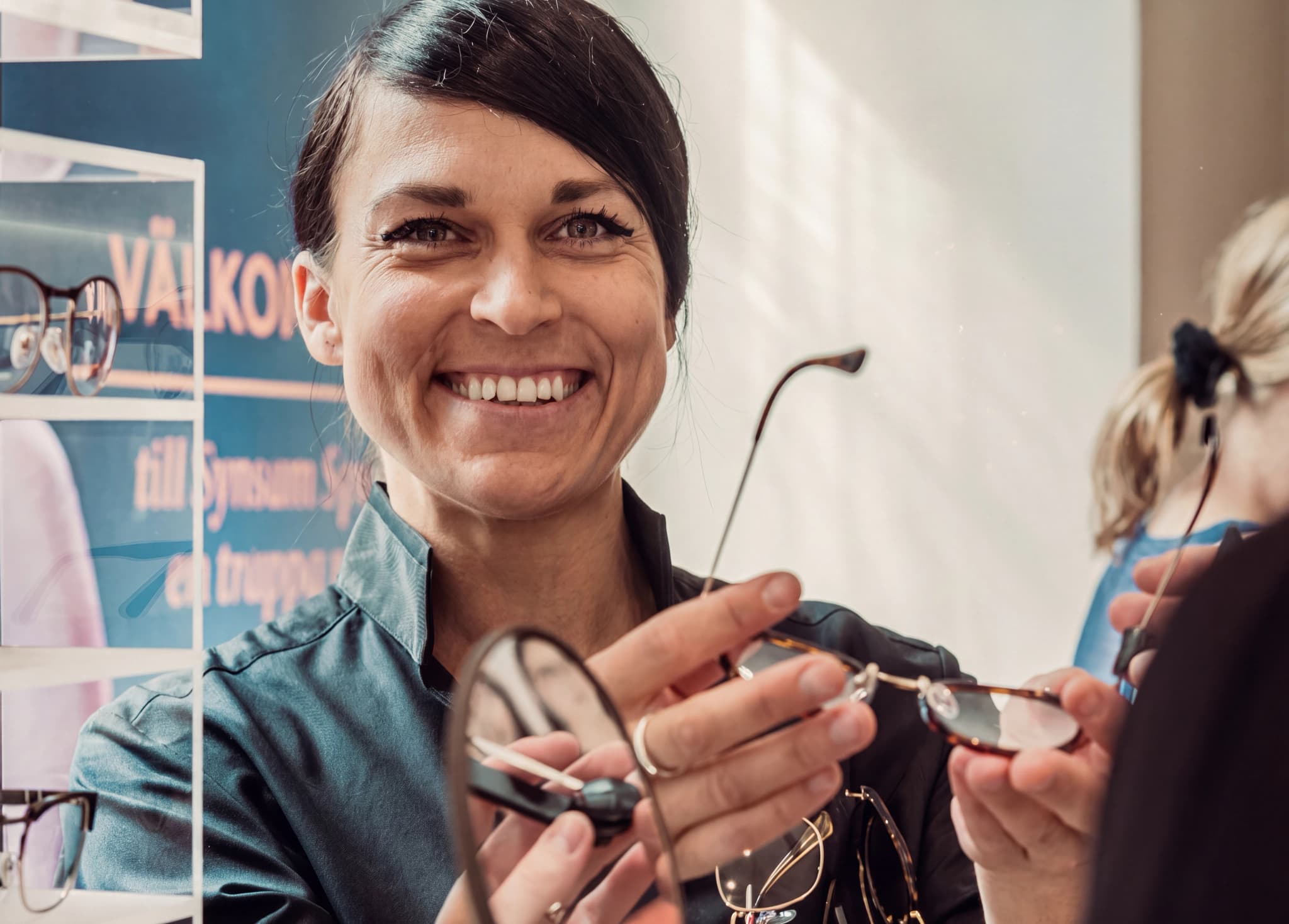 En optiker ler och håller upp ett par glasögon framför en kund i en butik.