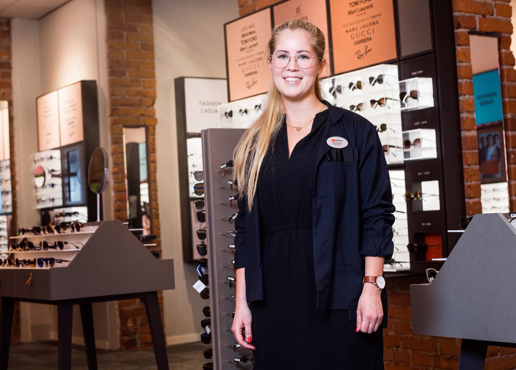 En ung kvinna med glasögon och en svart jacka står i en butik med glasögon i bakgrunden.